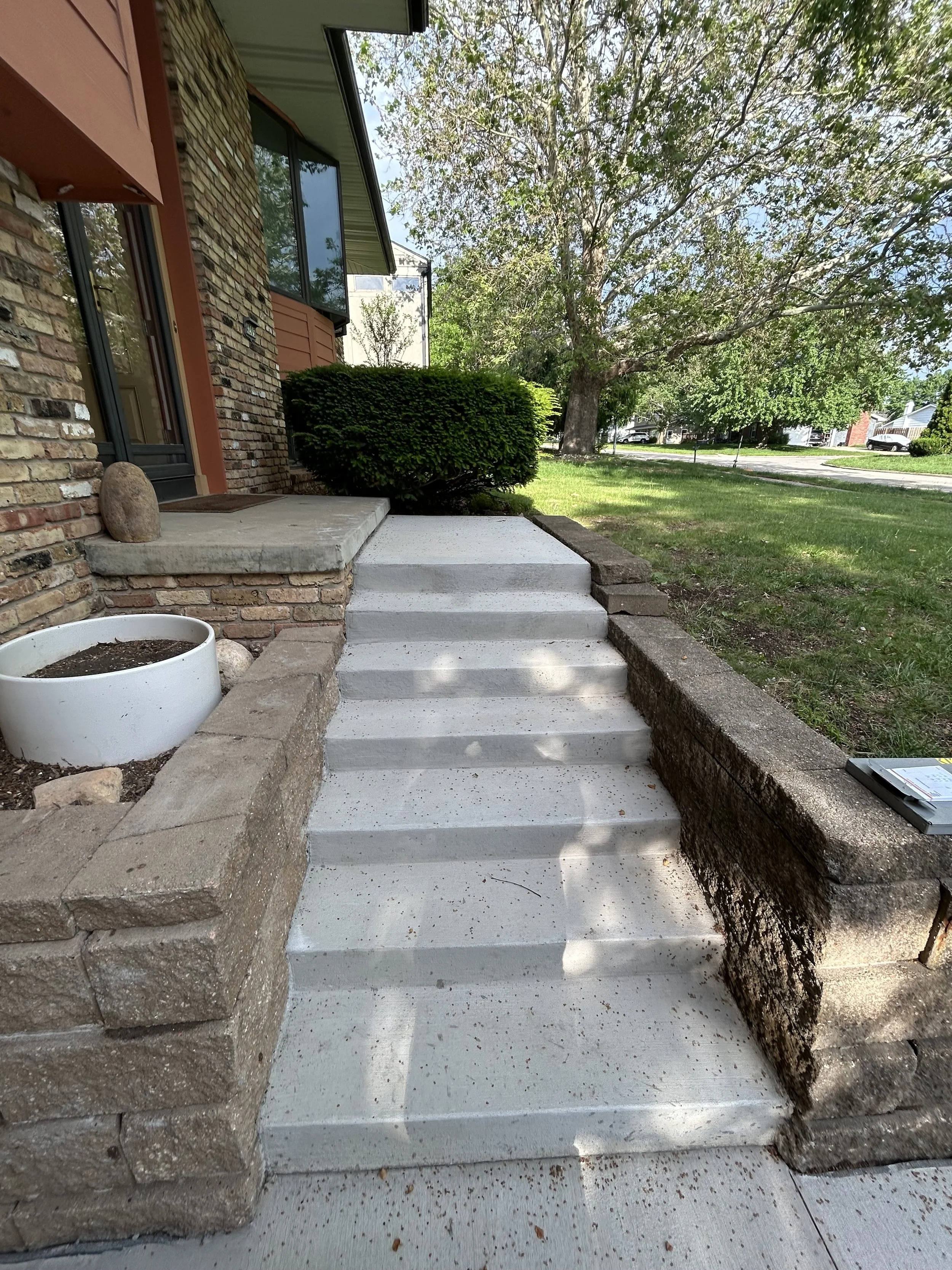 Concrete steps leading to a house front door, with a brick wall on the left and a garden with grass and trees on the right. A large tree with green leaves is visible in the background under a clear sky.