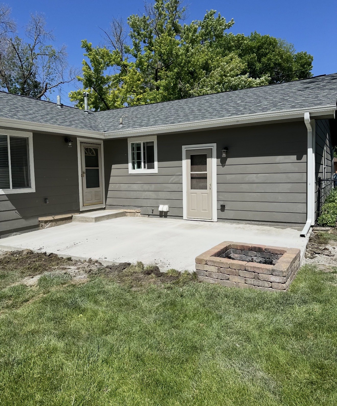 A backyard patio area with a concrete slab, a small brick fire pit, and a gray house with white trim and a sliding glass door.