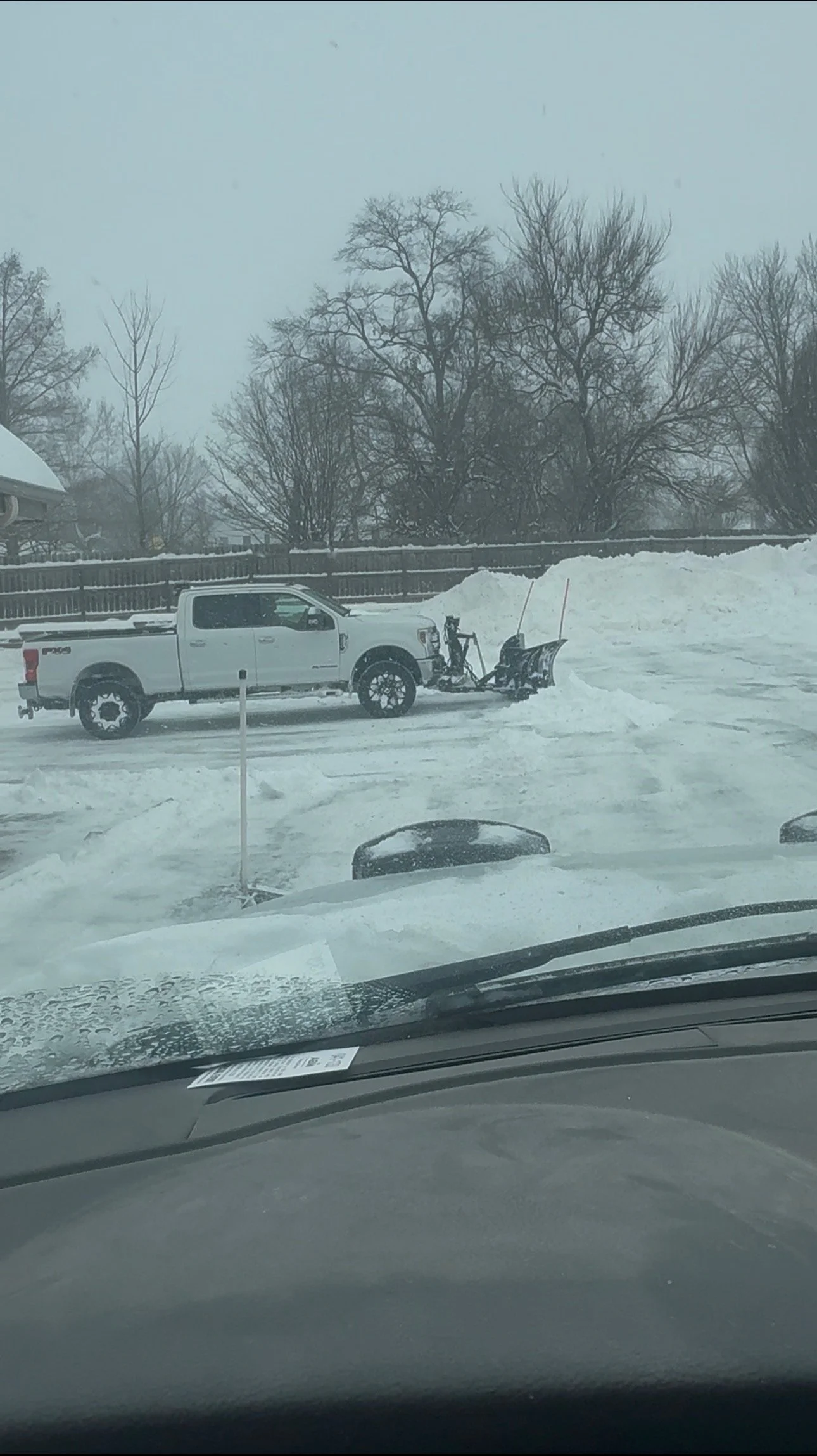 A white pickup truck parked on snow-covered ground with a snowplow attached to the front, and snow piles in the background, beneath leafless trees during a snowy day.