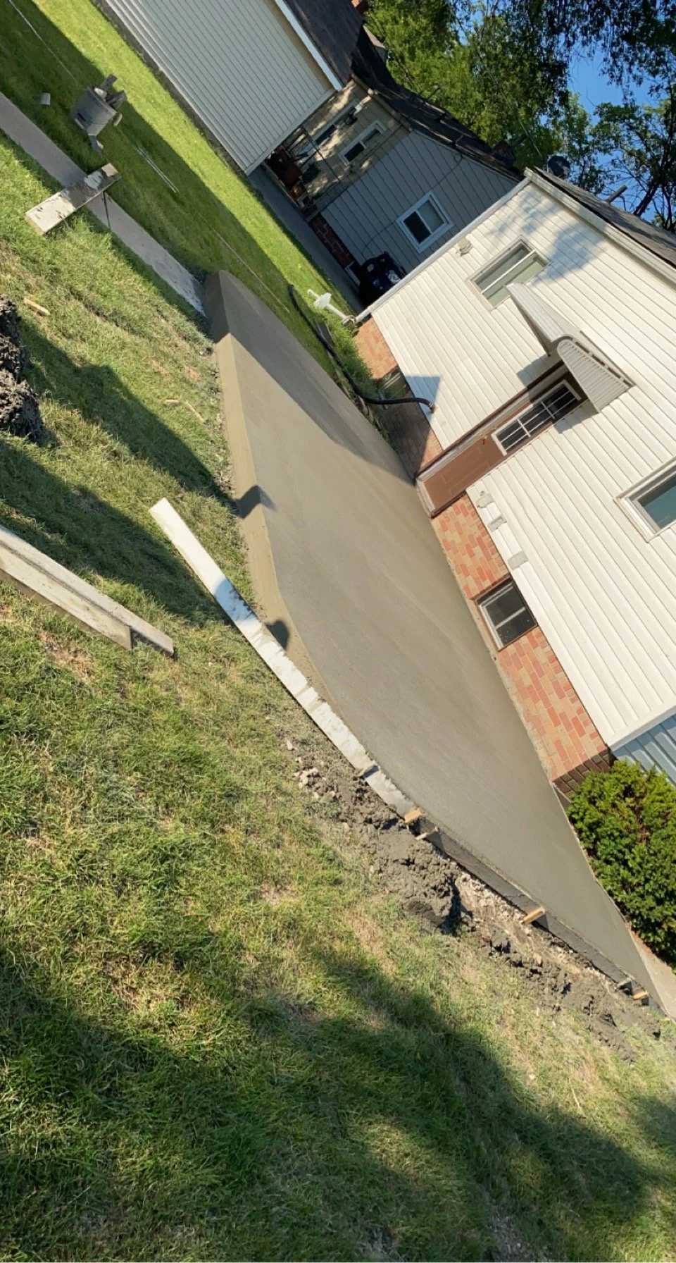 Newly poured concrete sidewalk in front of a house with white siding, a brown door, and windows. The sidewalk is still wet and drying, with construction materials around it.