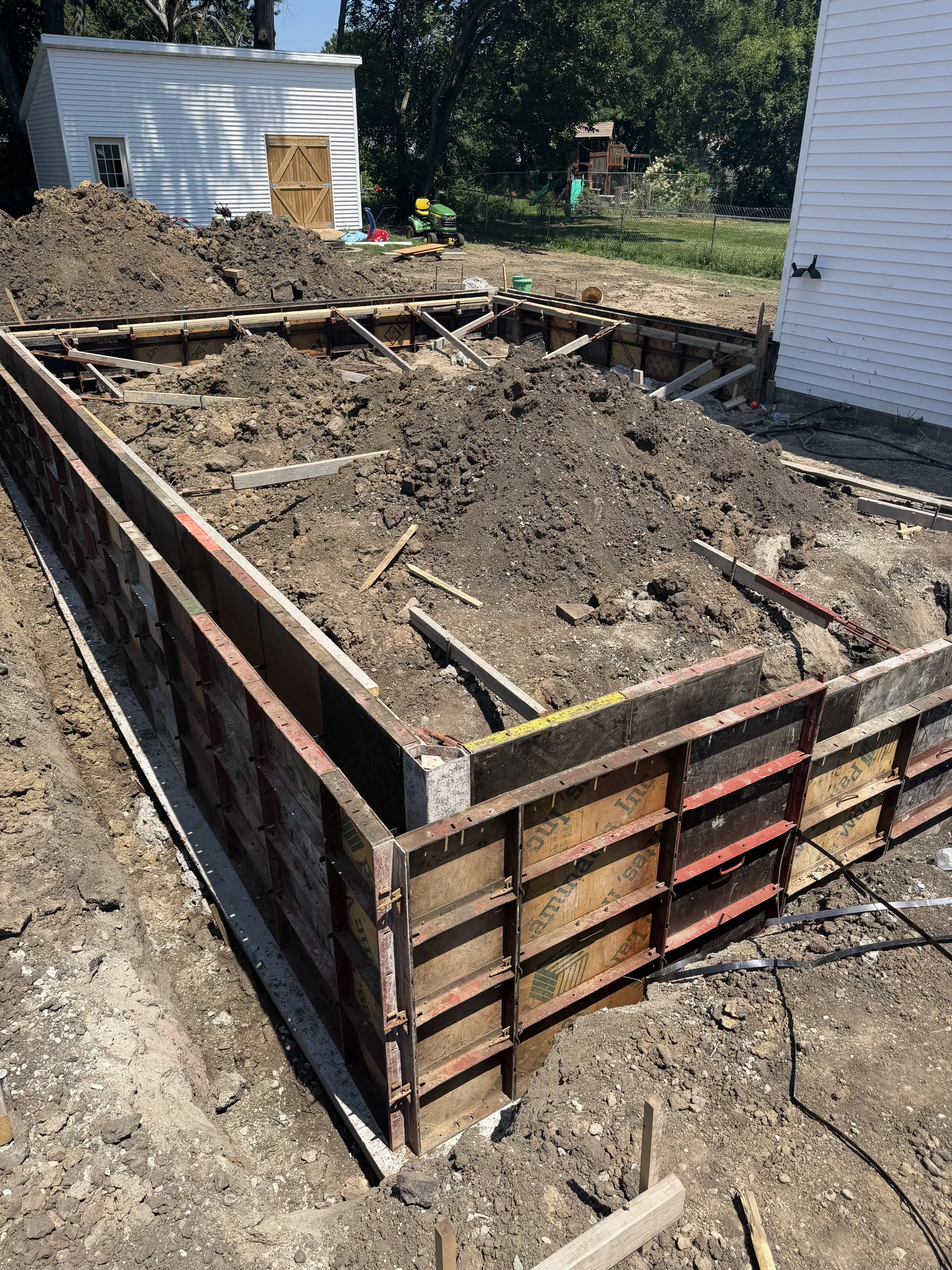 A construction site with wooden formwork for pouring concrete, surrounded by dirt and soil, in a backyard with a white shed and trees in the background.