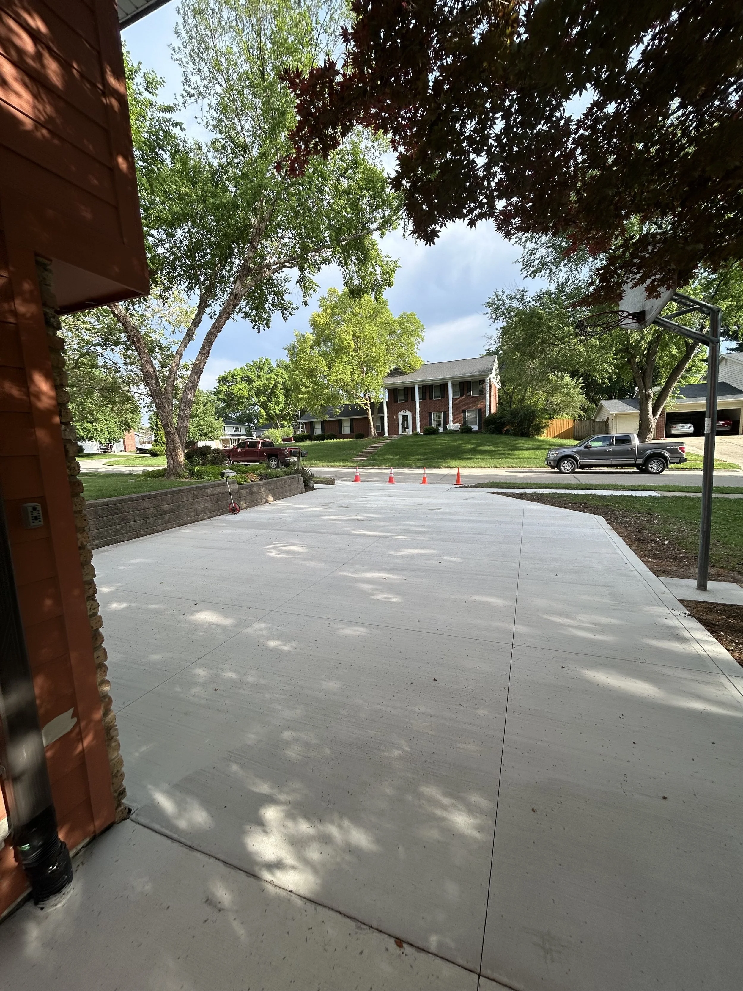 View of a suburban neighborhood with a concrete driveway, a basketball hoop on the right, parked cars, green trees, and houses in the background under a partly cloudy sky.