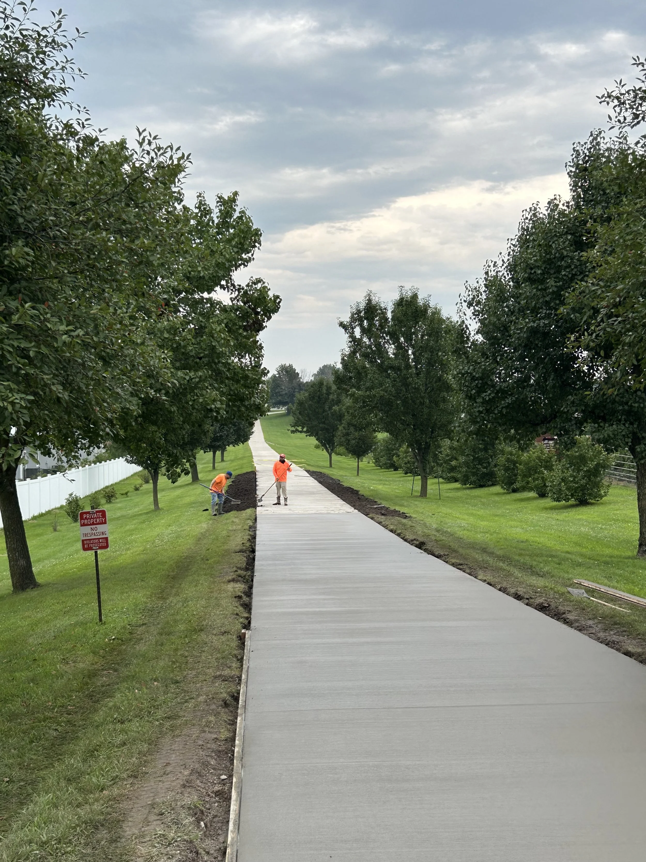 Two construction workers in orange shirts and hats laying a new concrete sidewalk on a grassy area, with trees and a cloudy sky in the background.