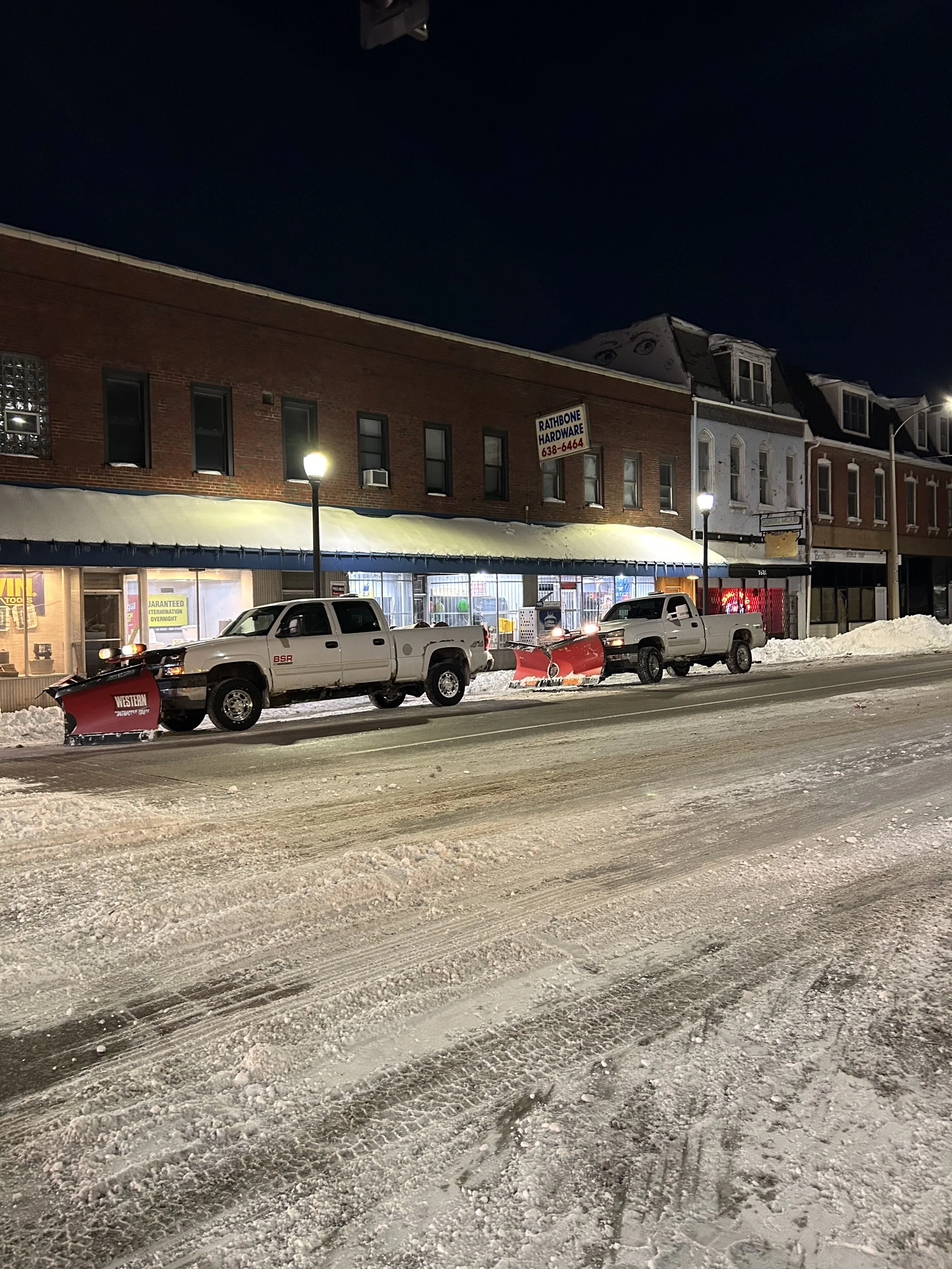 Street scene at night with snow-covered ground, parked trucks, a storefront with large windows, and buildings with brick and wood facades illuminated by streetlights.