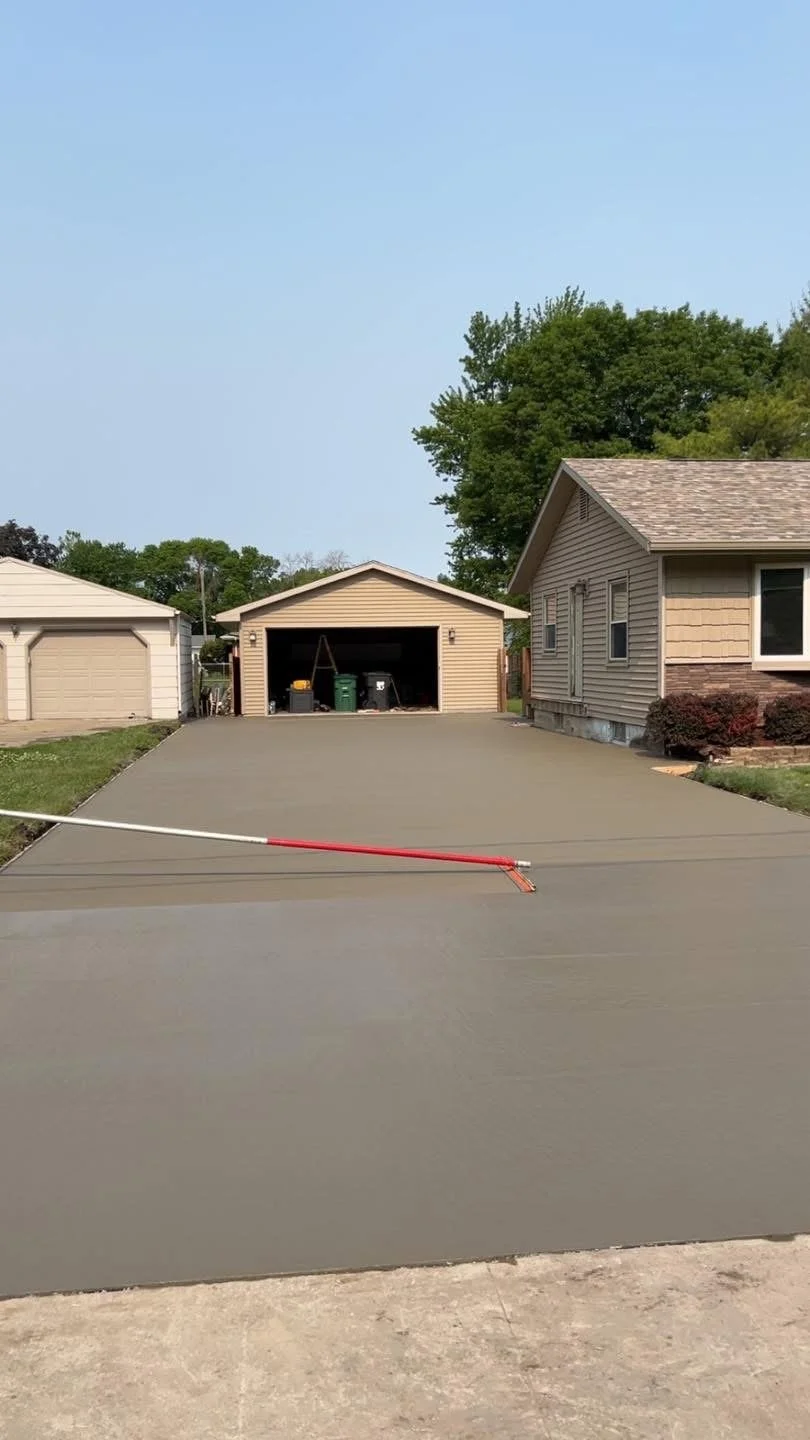 Recently poured concrete driveway in front of a garage, with a red and white leveling tool laying across it. House and trees in the background.