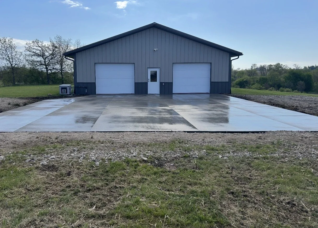 A gray metal building with two large garage doors, a smaller door with a window, and a concrete driveway in front. The area around the driveway is grassy with some patches of dirt, and there are trees in the background under a blue sky.