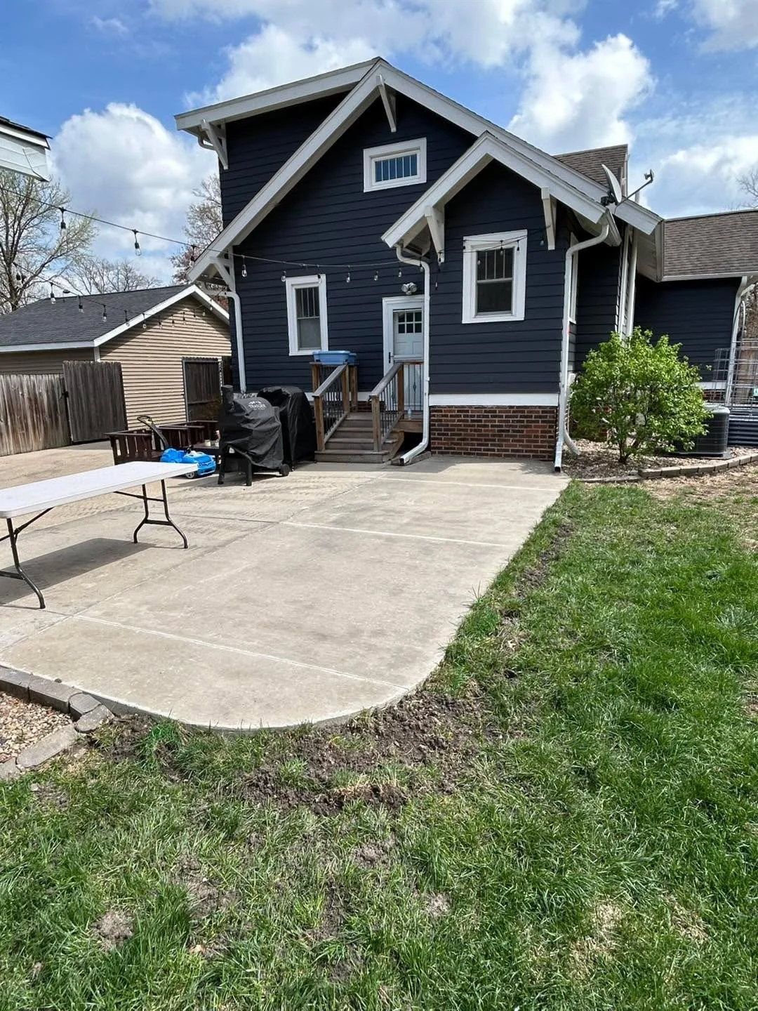 Backyard with a concrete patio, a small porch with stairs, and a siding house painted dark blue with white trim, surrounded by a wooden fence and green grass, under a partly cloudy sky.