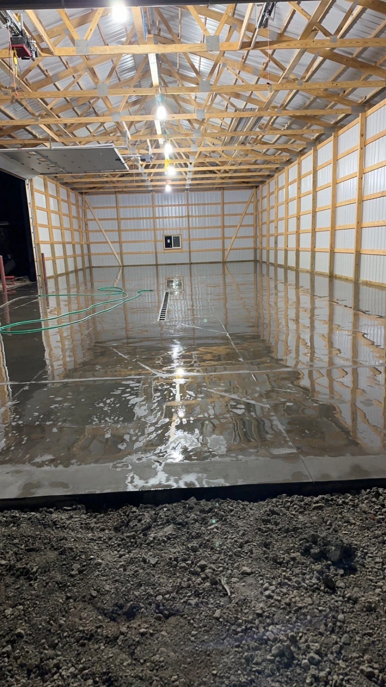 An unfinished building with a wooden framework and a concrete floor that has been recently poured, still wet, with foam soap on the surface, and gravel at the edges.