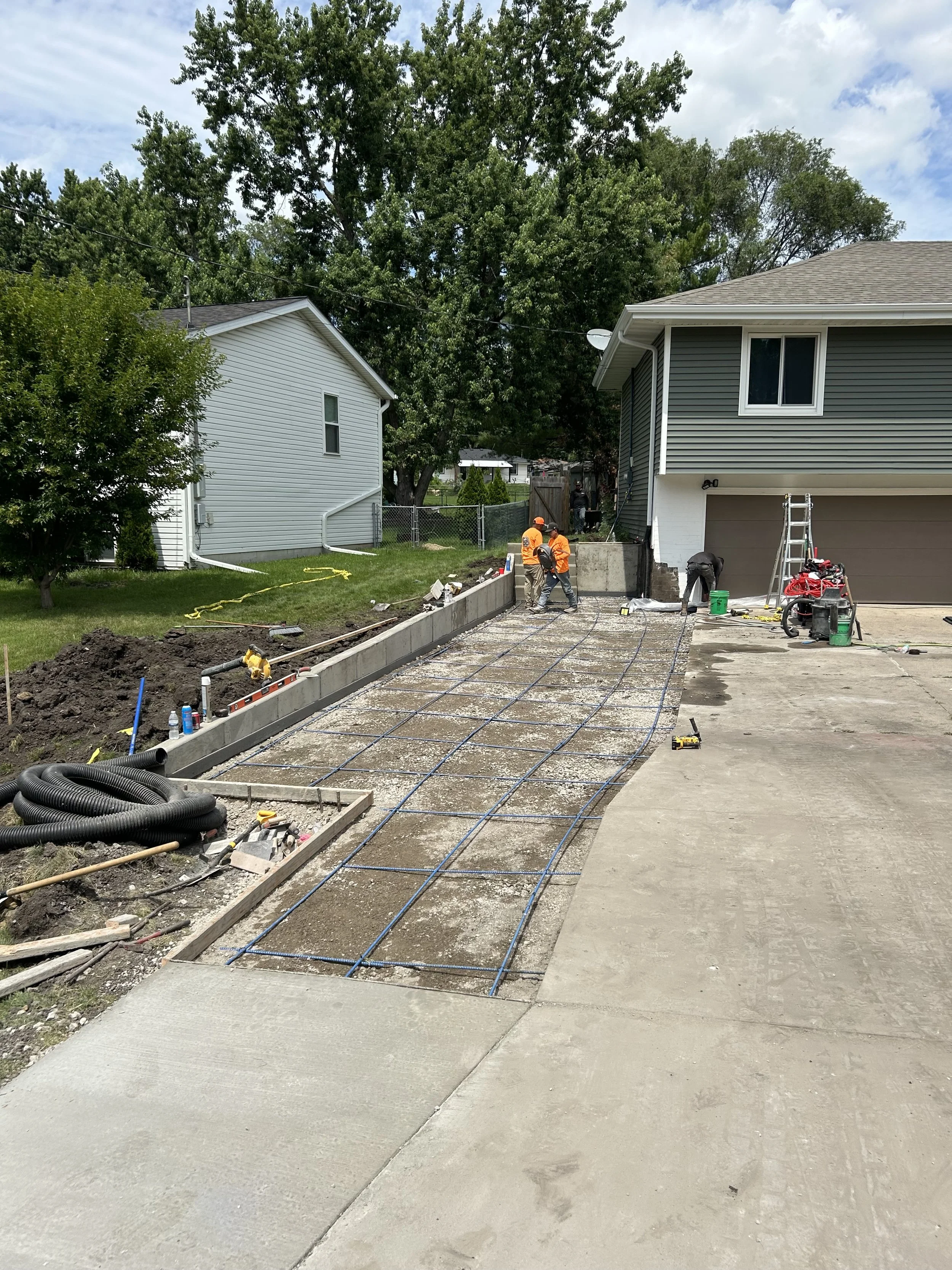 Construction workers installing a concrete driveway, with rebar and construction tools visible, in a residential neighborhood with houses, trees, and a chain-link fence.