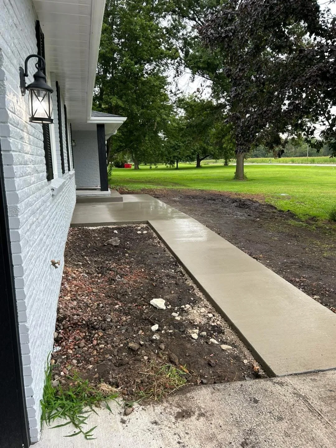 Newly poured concrete sidewalk next to a white brick house with black window shutters and a black wall-mounted lantern, on a cloudy day with green trees and a grassy field in the background.