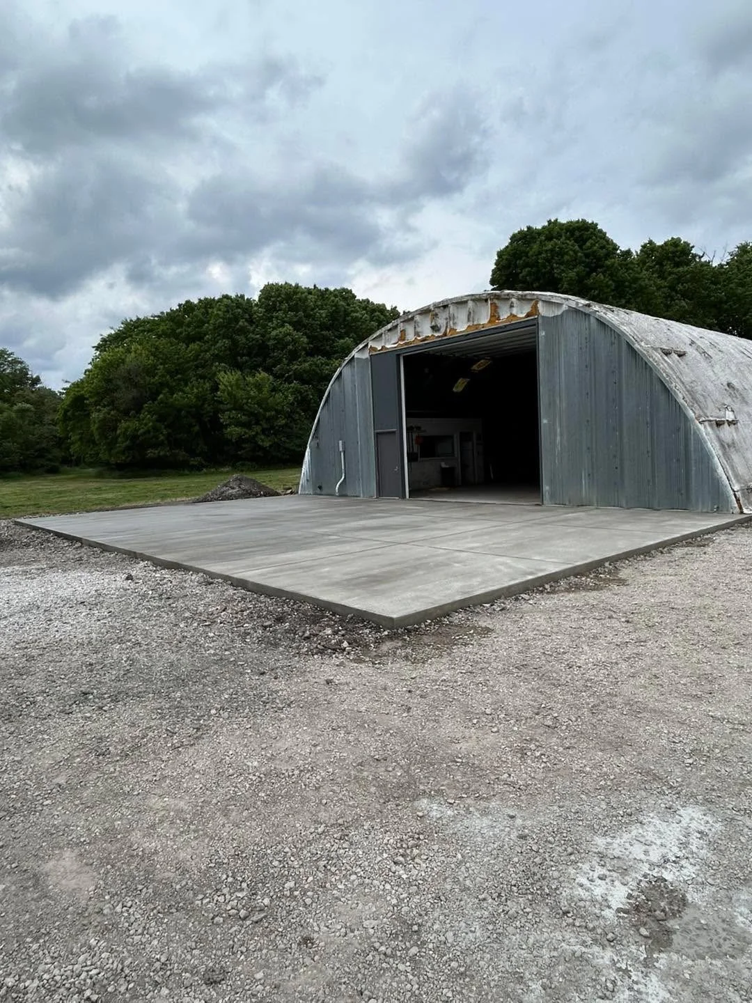 A metal Quonset hut with a concrete pad in front of it, located outdoors under a cloudy sky, surrounded by trees and gravel ground.