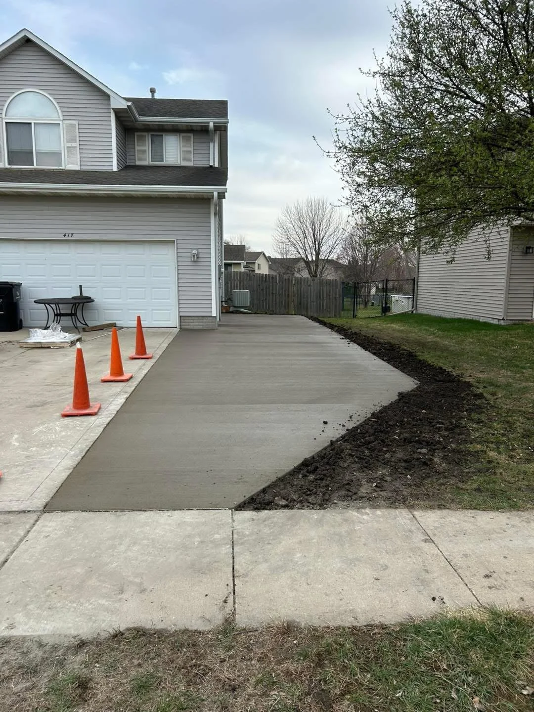 Freshly poured concrete driveway on a suburban house with orange traffic cones and a partially paved sidewalk, with grass and trees in the background.