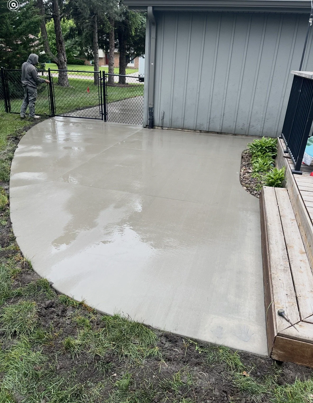 A concrete patio area next to a house with a wooden deck and a black metal fence gate being closed by a person dressed in gray. The patio is wet, likely from recent rain, and is bordered by grass and flower beds.