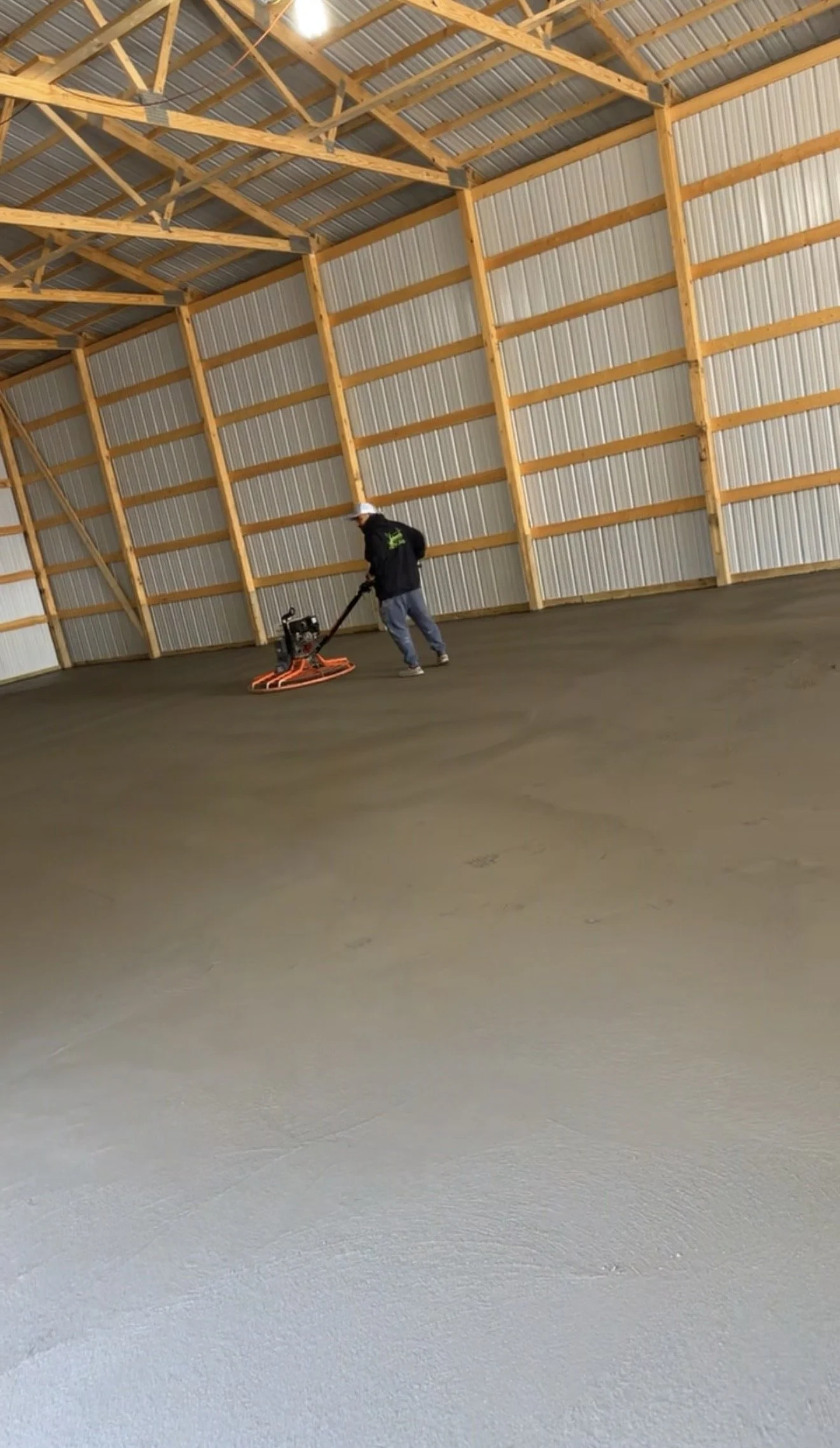 A person operating a floor grinder or polisher inside a large wooden barn with metal roofing, preparing the concrete floor.