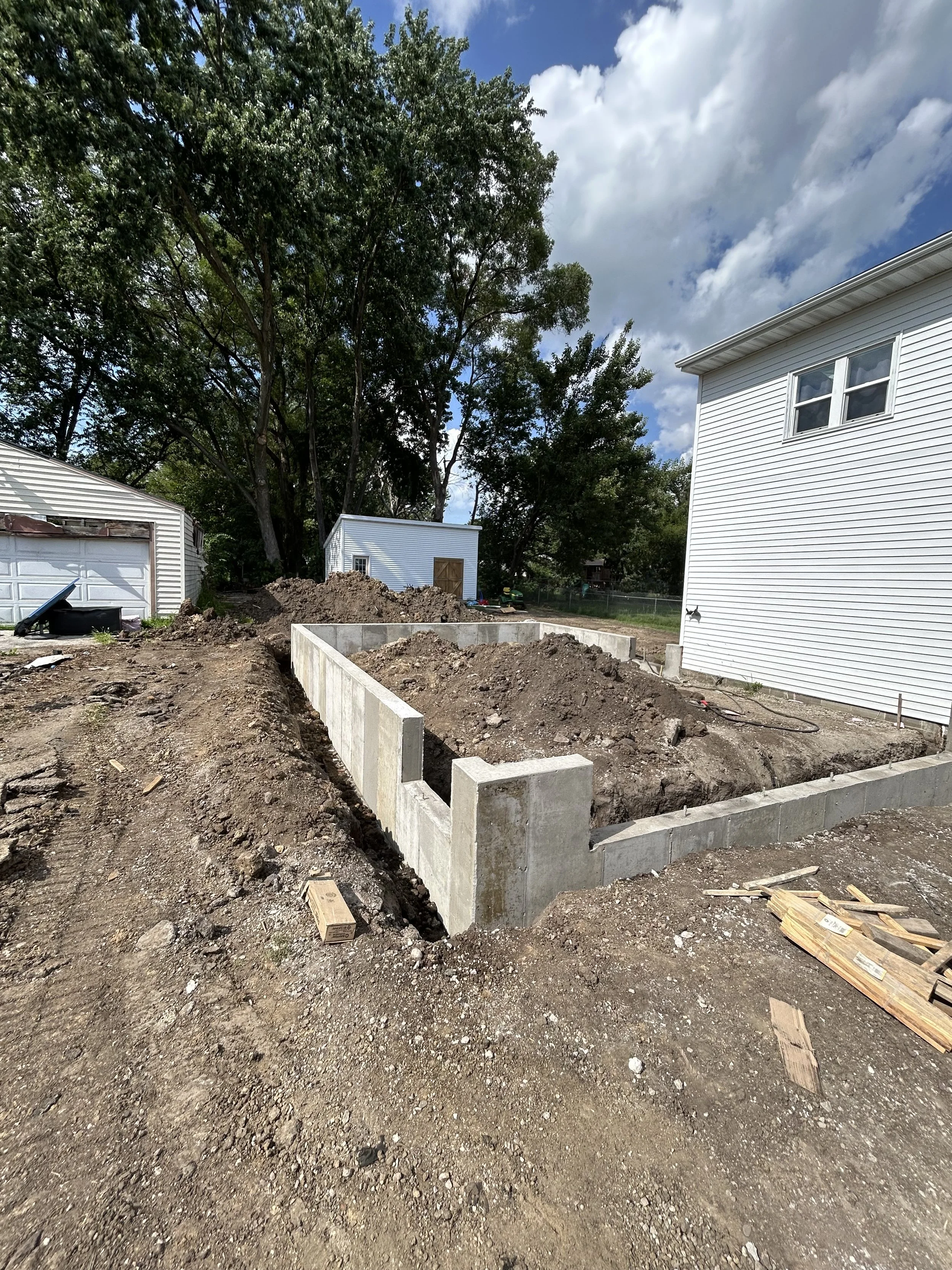 Front yard with dirt and construction foundation, two-story white house on right side, white shed with brown door in background, large trees with green leaves, cloudy sky.