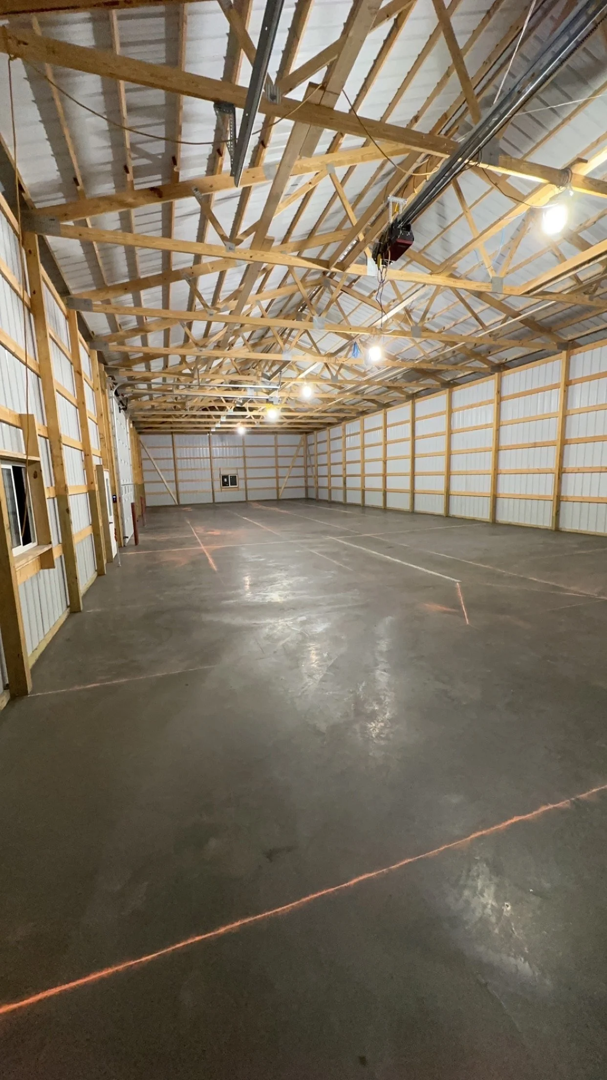Empty indoor space with a concrete floor, wooden framing, and metal roofing, illuminated by ceiling lights.