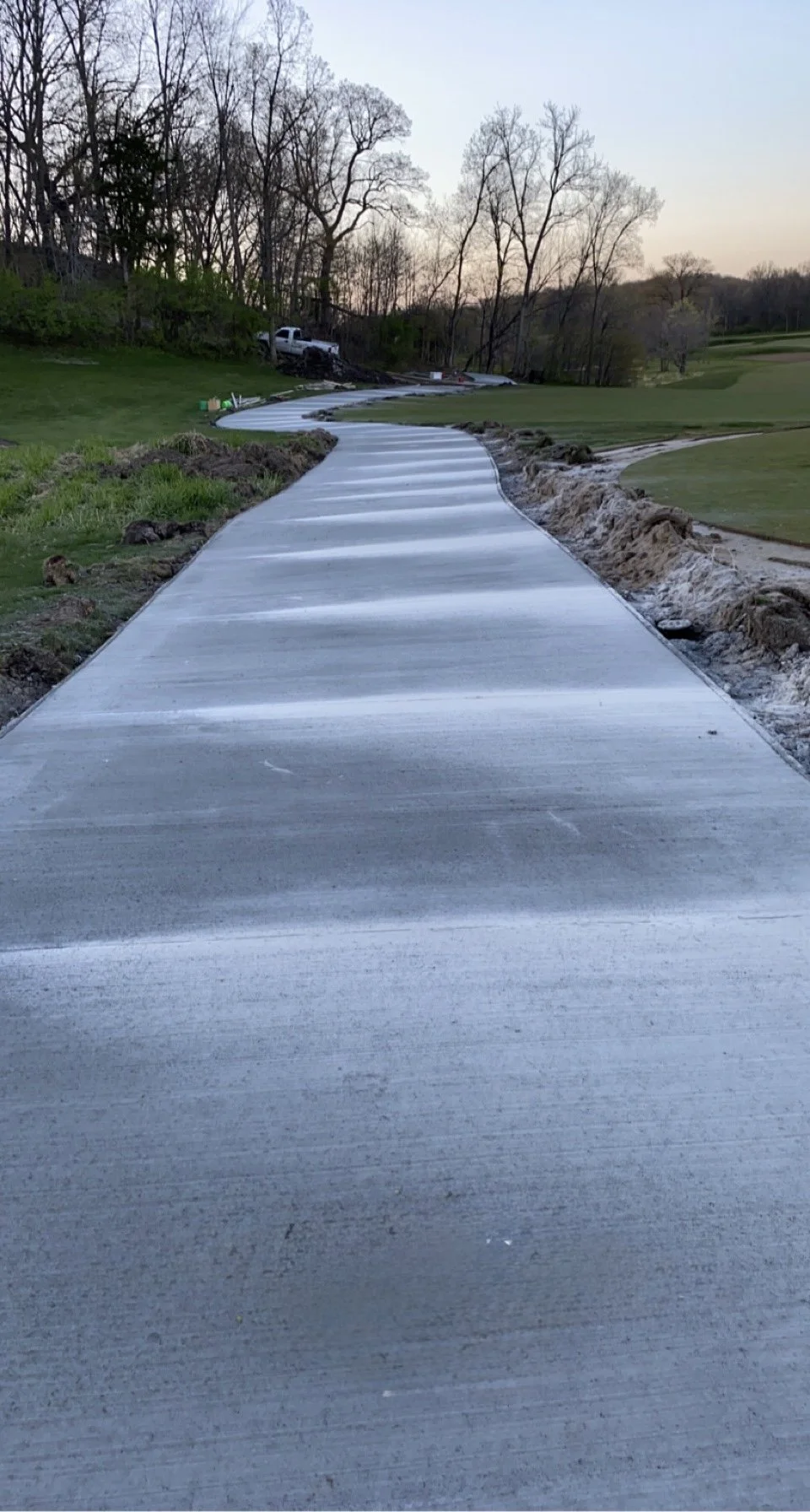 A newly constructed concrete sidewalk winding through a grassy area with a slight curve, bordered by dirt and soil, with trees and a truck in the background under a clear sky.