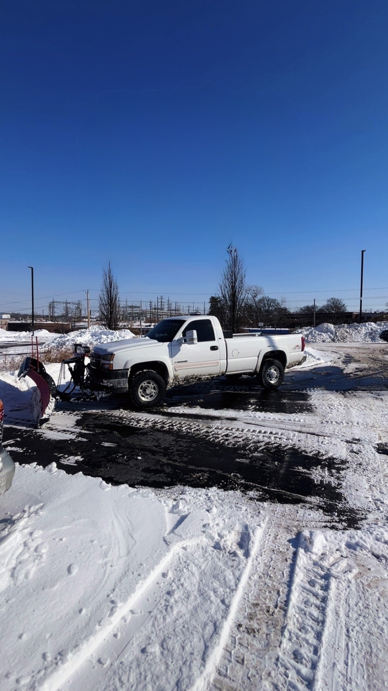 White pickup truck parked in a snowy lot with snow removal equipment attached to the front, under a clear blue sky, with snow piles and utility poles in the background.