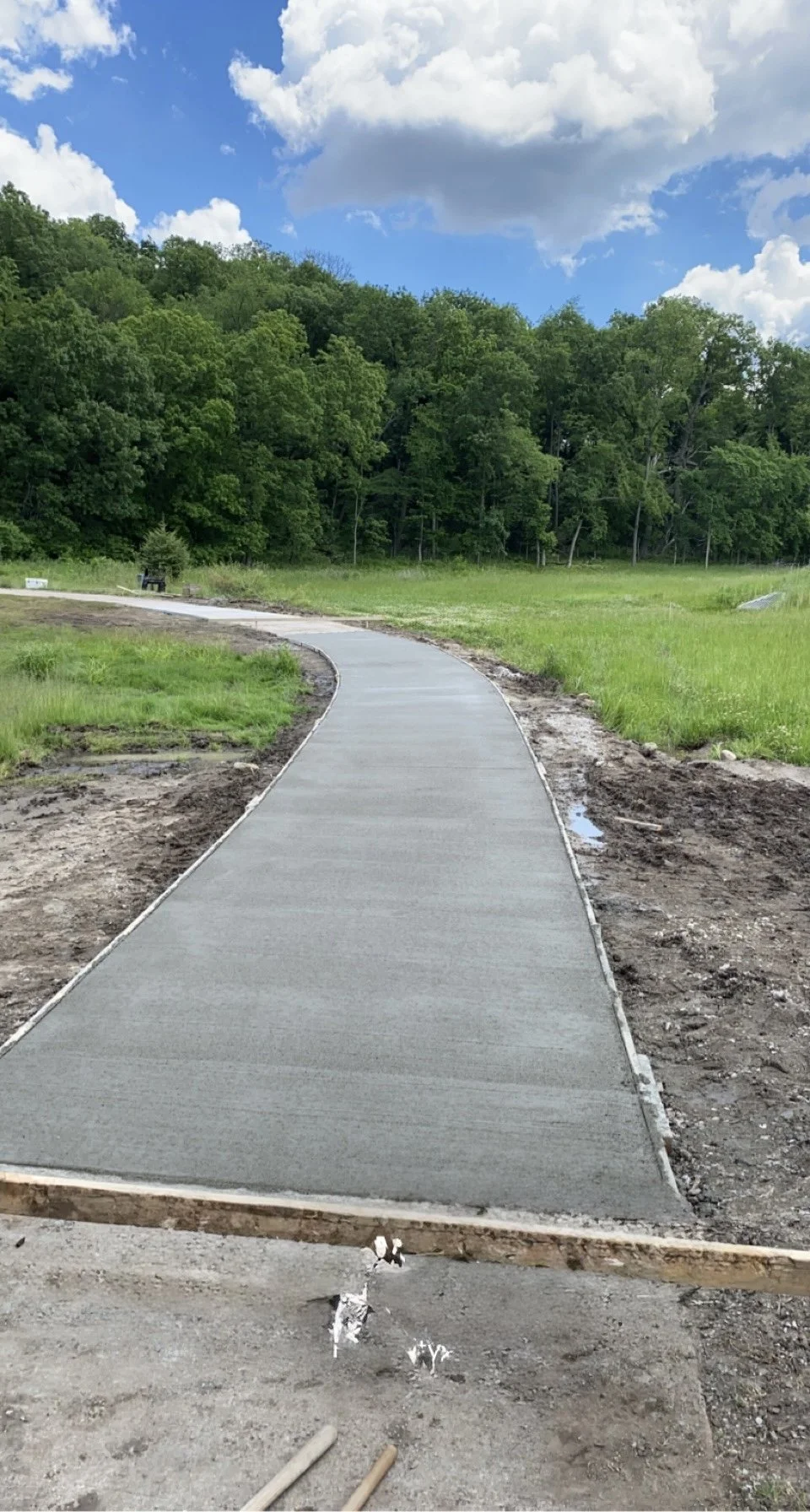 Newly constructed concrete sidewalk winding through a grassy area near a wooded landscape with green trees and blue sky.