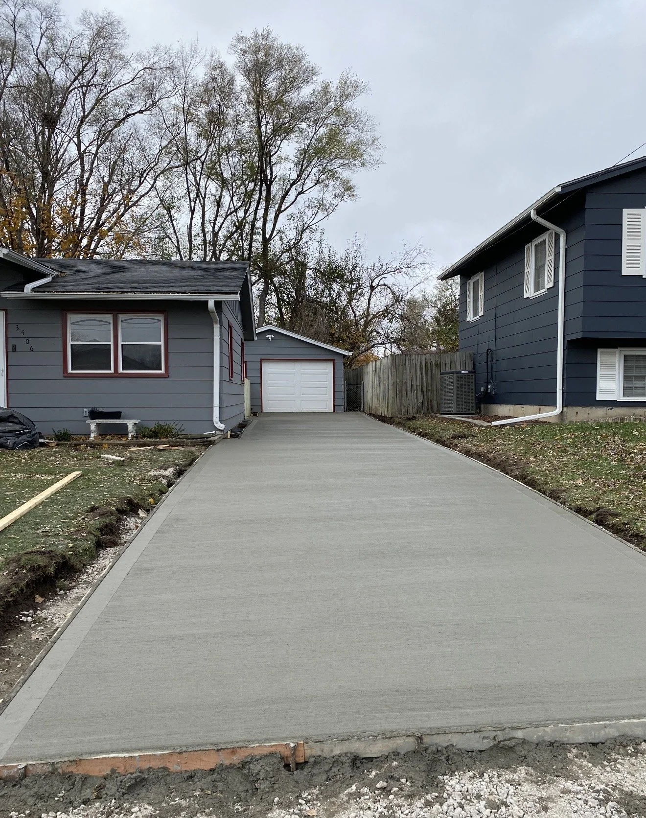 View of a newly paved concrete driveway leading to a garage, between two houses with blue siding, in a residential neighborhood. Trees are visible in the background.