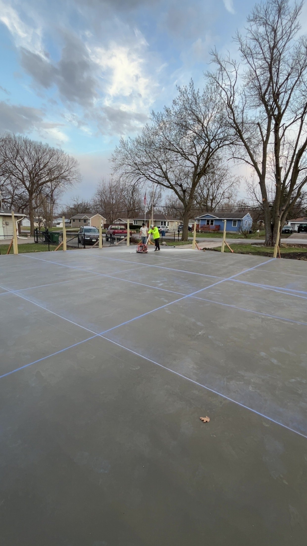 Two workers are marking a concrete surface with lines, likely for a new construction or sports court, in a residential neighborhood with trees and houses in the background.