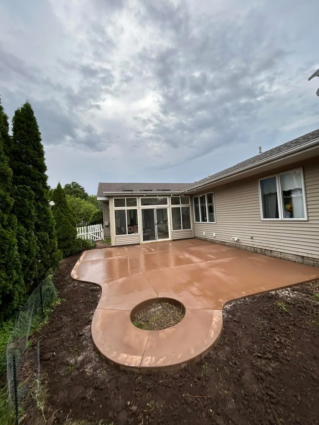 Recently poured concrete patio in backyard with curved design and a circular cutout, surrounding a dirt area, adjacent to house with beige siding and multiple windows, overcast sky.
