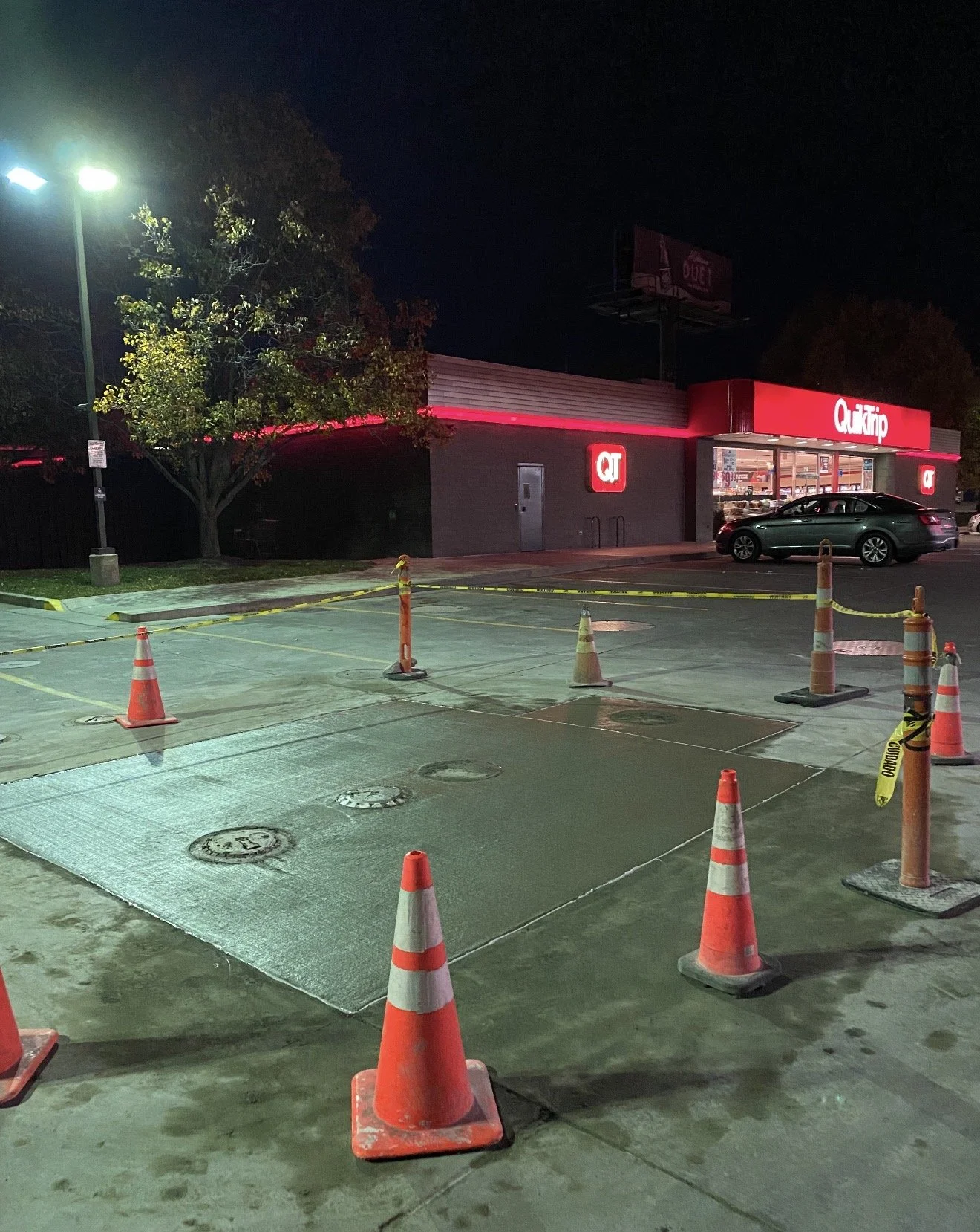 Empty parking lot with orange traffic cones and yellow caution tape, and a QT convenience store in the background at night.