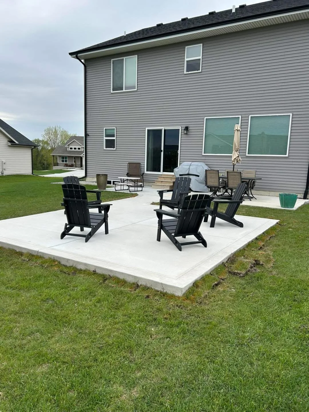 Backyard patio with black Adirondack chairs, patio table with umbrellas, grill covered with a tarp, and steps leading to the house with gray siding and sliding glass door.