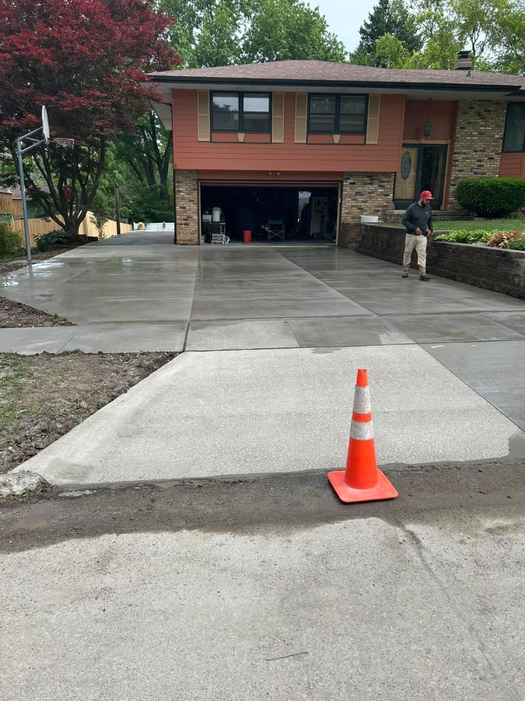 Recently poured concrete driveway leading to a house, with a construction cone placed at the front. A worker in a black hoodie and red helmet is walking on the newly finished driveway, which leads to an open garage. The house features a brick and woo