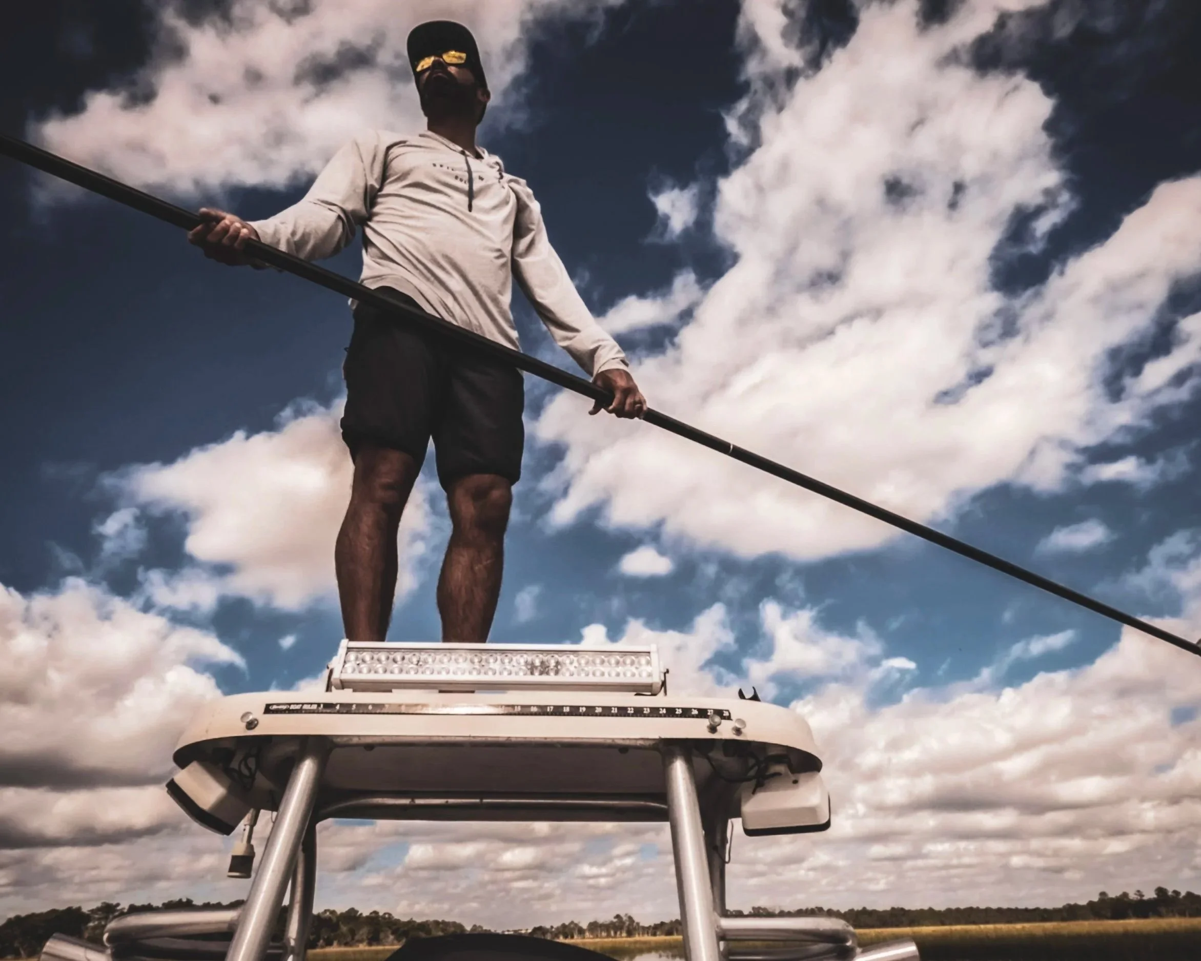 A person wearing a baseball cap and sunglasses standing on a boat or dock, holding a pole, with a cloudy sky in the background.