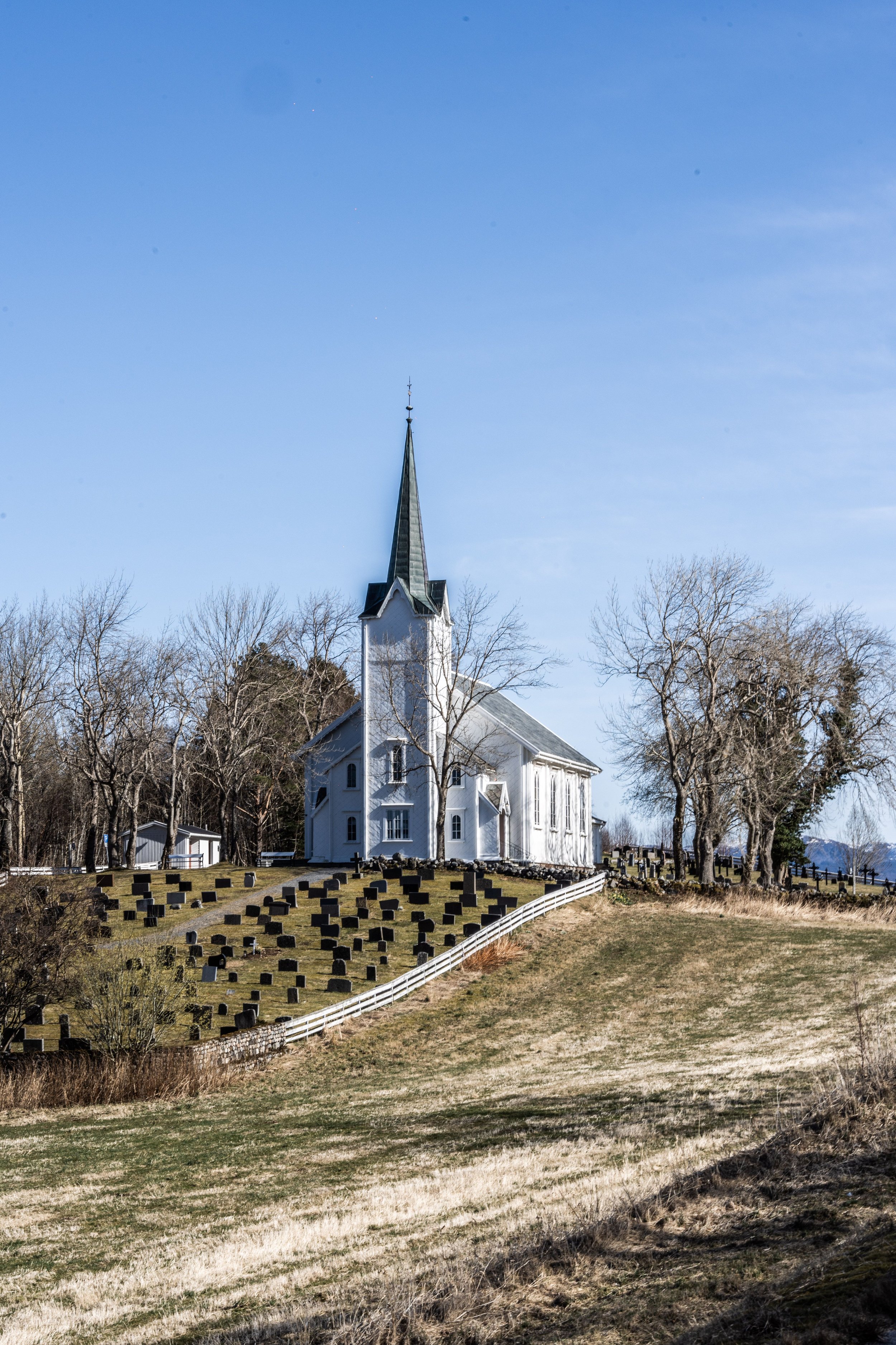 A white church with a tall, pointed steeple and graveyard in front, surrounded by leafless trees under a clear blue sky.
