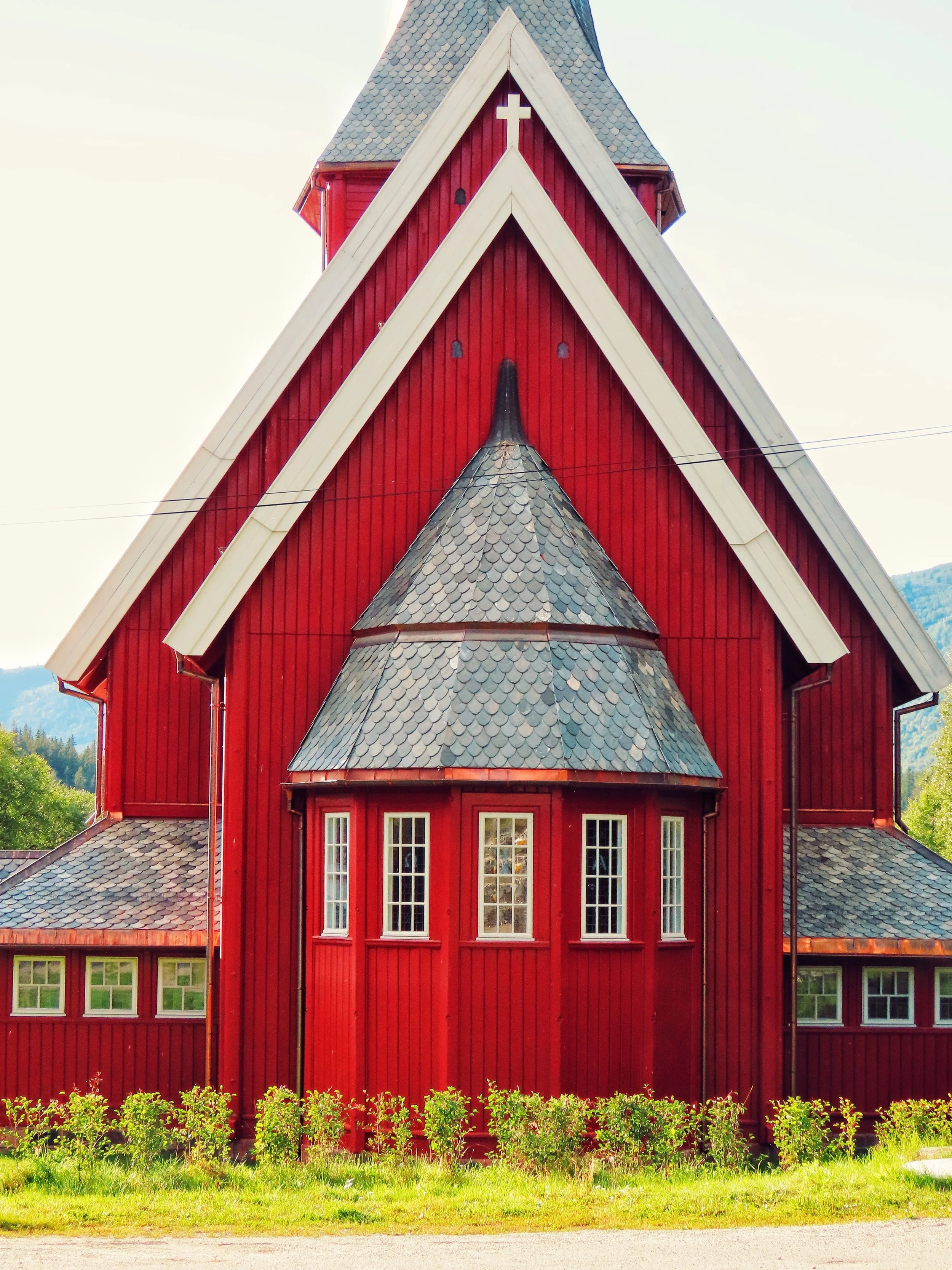 Front view of a red wooden church with white trim, multiple windows, and a steep roof topped with a cross, set against a backdrop of trees and mountains.