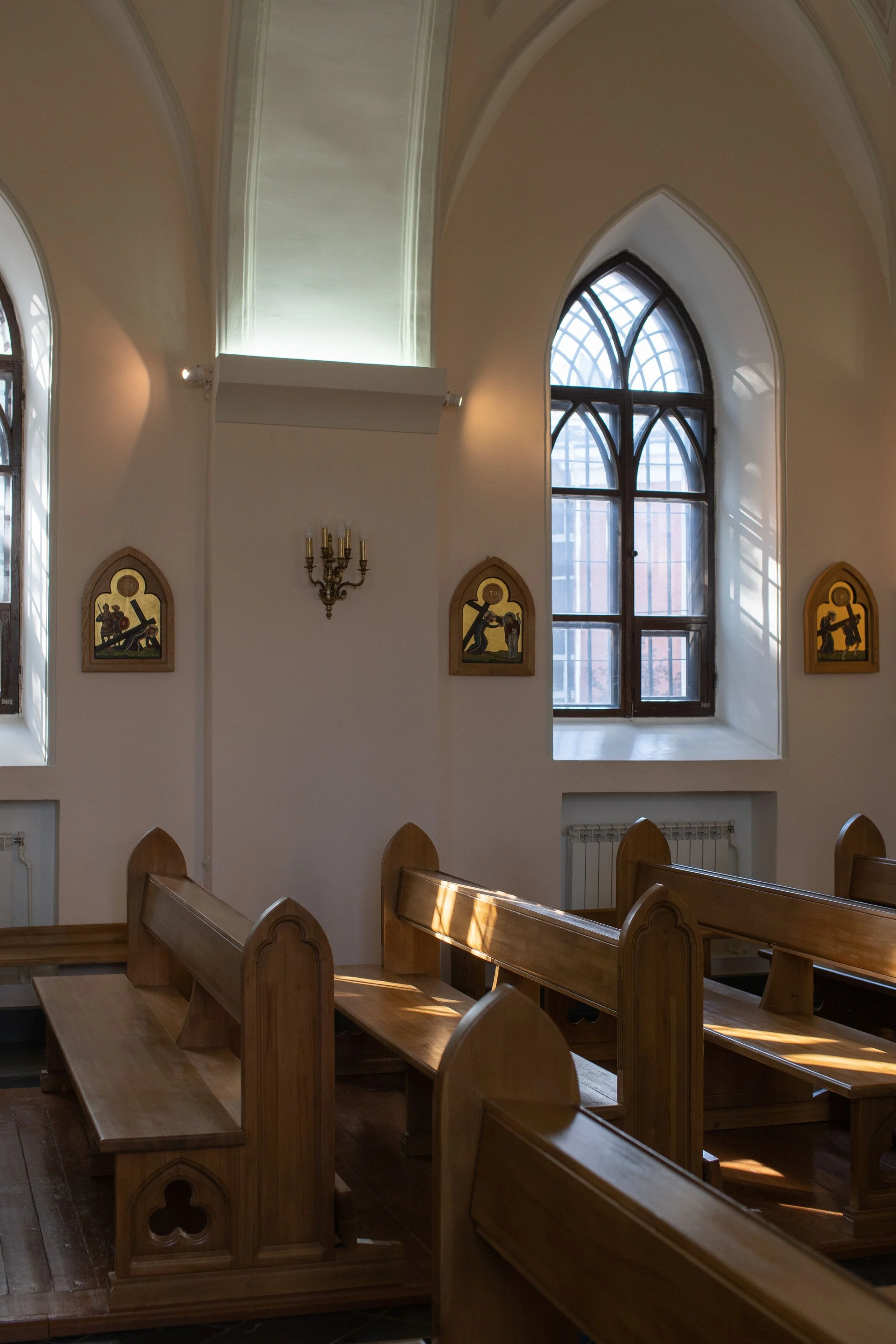 Inside a church with wooden pews, large arched windows allowing natural light, religious artwork on the walls, and a candelabra on the wall.