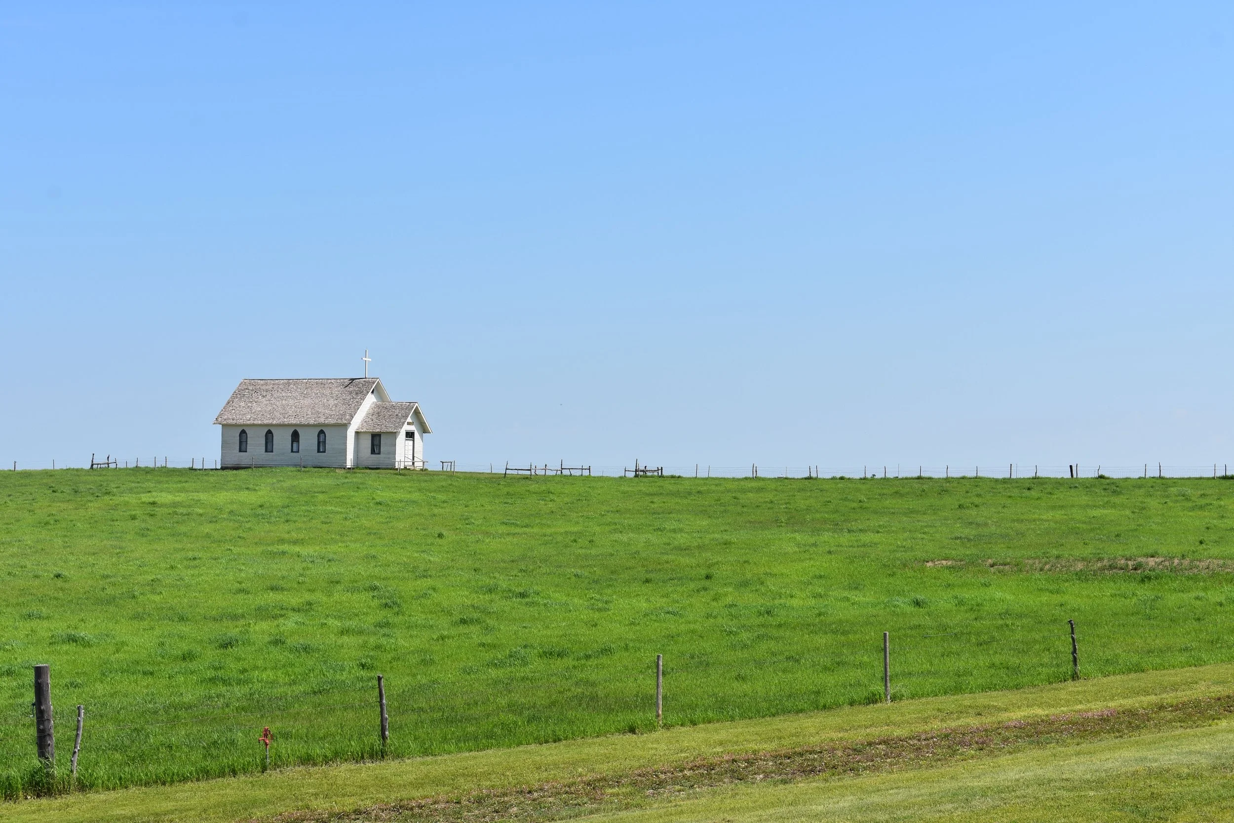 A small white church with a cross on the roof, situated in a lush green field under a clear blue sky.