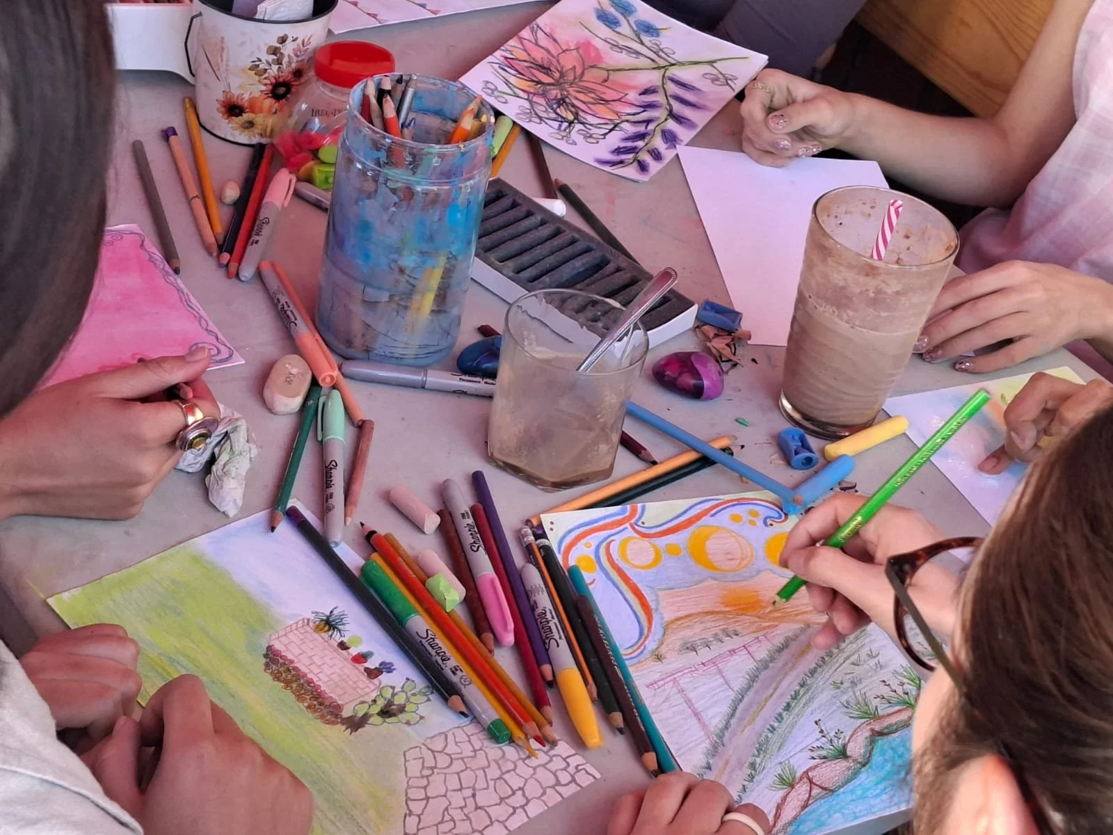 Grupo de personas pintando y dibujando en una mesa con papeles, marcadores, colores y utensilios de arte. Hay bebidas en vasos con popotes y algunos objetos decorativos en la mesa.