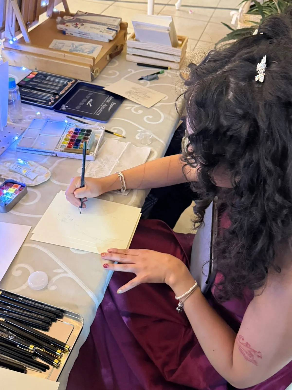 Una mujer con cabello rizado y con accesorios en el cabello está haciendo ilustraciones o dibujos en un cuaderno sobre mesa decorada con mantel con patrones blancos. En la mesa hay varios artículos de arte, incluyendo acuarelas, plumas y papel, así como un tabique de madera con libretas y bolígrafos.