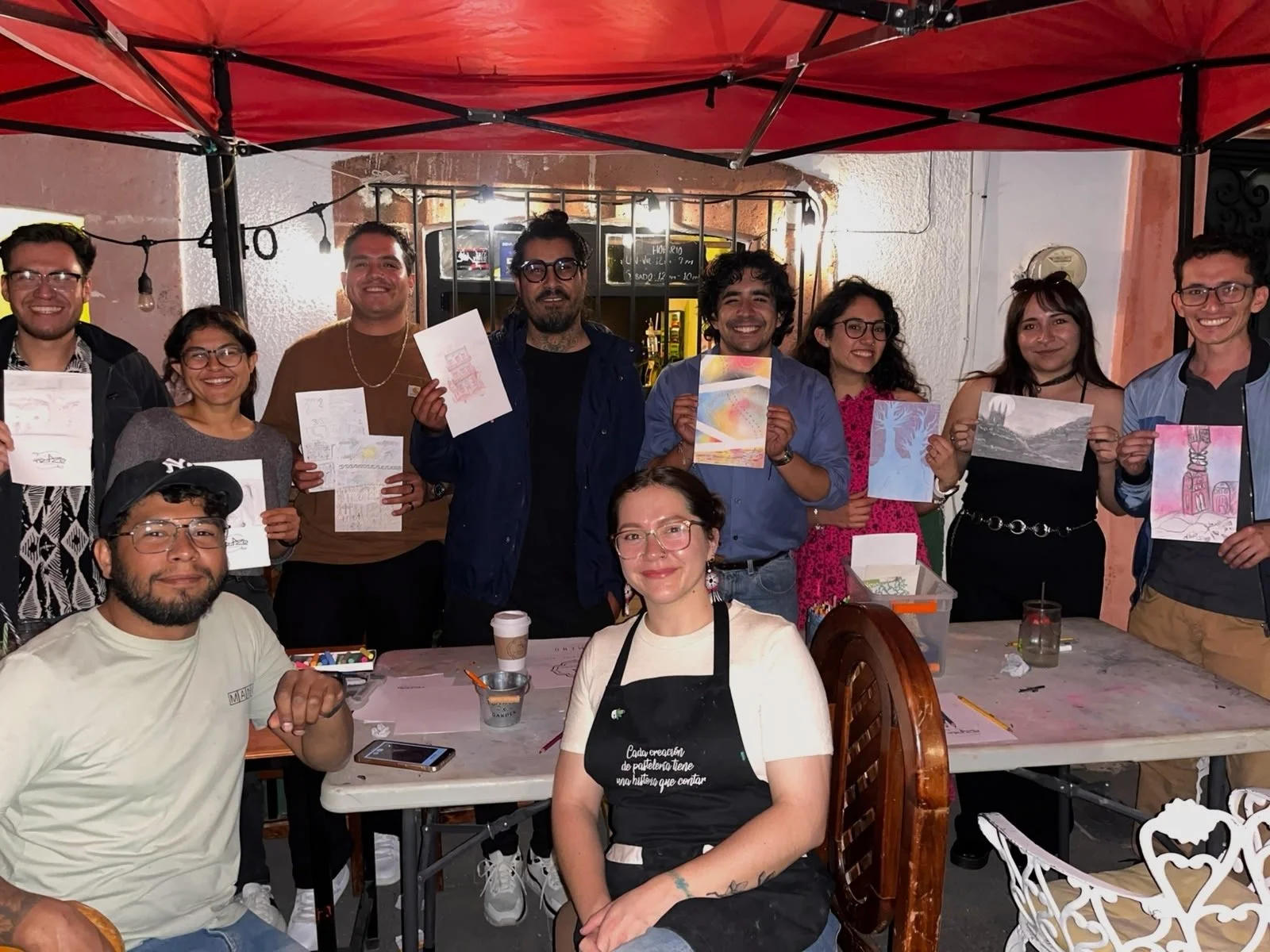 Grupo de personas en una reunión, algunas sosteniendo dibujos y obras de arte, en un ambiente con toldo rojo y decoración de fiesta.