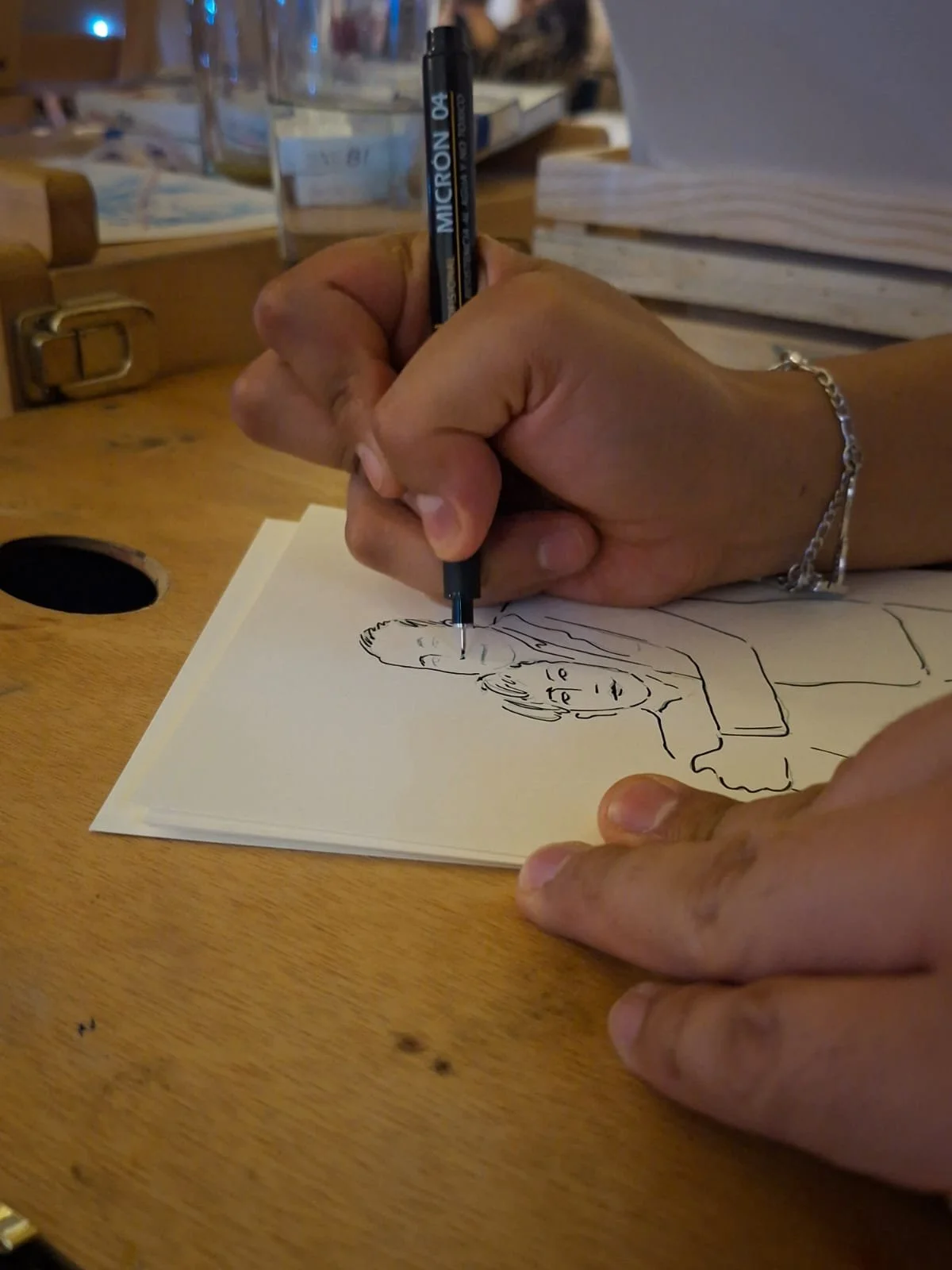 Una persona dibuja una caricatura de una mujer en una hoja de papel, usando un bolígrafo. La escena se desarrolla en un entorno que parece un café.