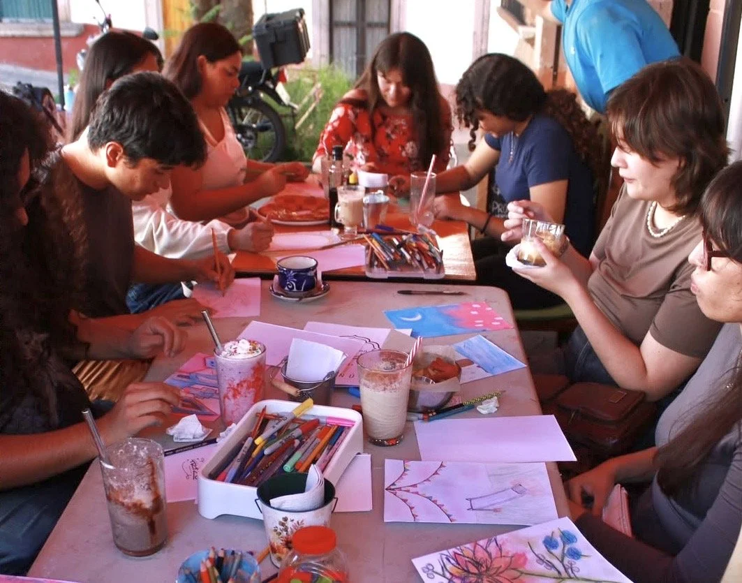 Un grupo de personas sentadas alrededor de una mesa participando en una actividad artística, con papeles y lápices, en un ambiente alegre y creativo.