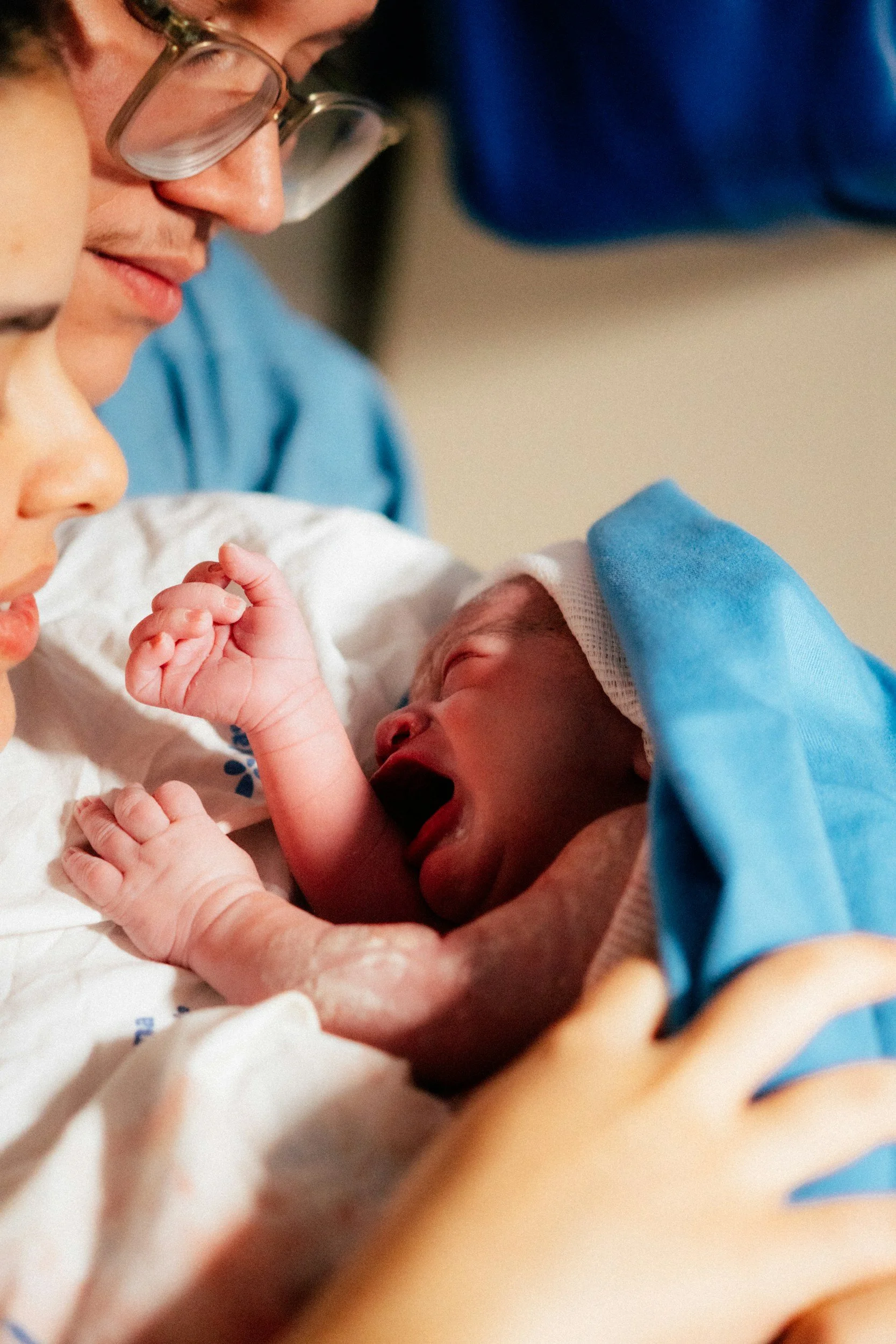 A newborn baby lying on a hospital bed with a nurse in blue scrubs and glasses attending to them. The baby has a hand raised near the face and appears to be crying or yawning.
