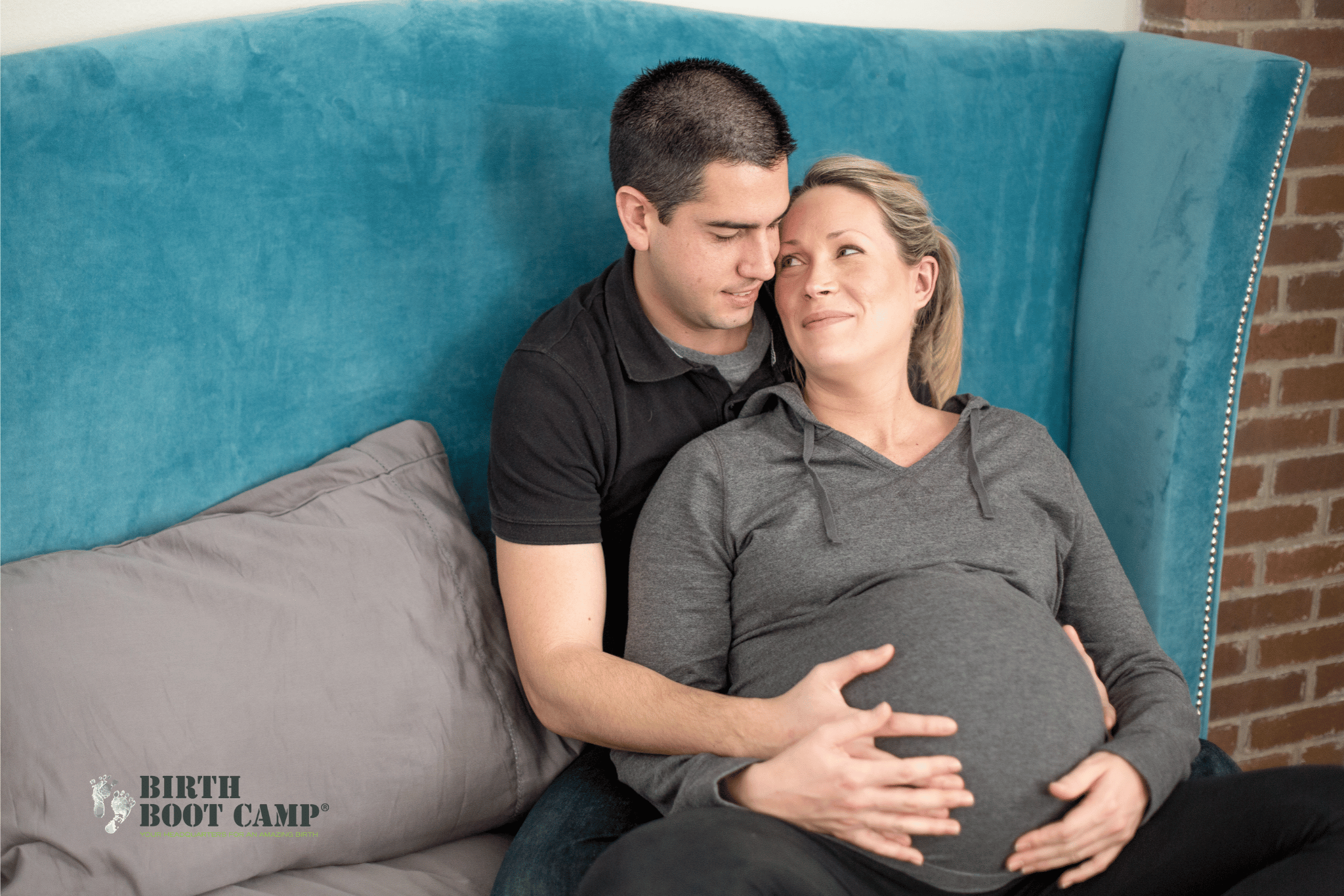 A man and a pregnant woman sit on a bed, lovingly touching her belly, with a blue headboard and brick wall in the background.