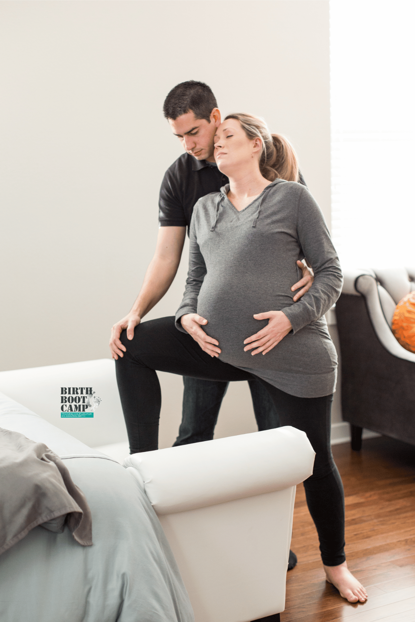 A pregnant woman with closed eyes leaning back against a man who is standing behind her. The man is supporting her by holding her. They are in a bright room, possibly during a childbirth class, with a beige couch and some clothing on a white armchair visible. There is a logo at the bottom left corner that reads 'Birth Boot Camp' with additional text underneath.