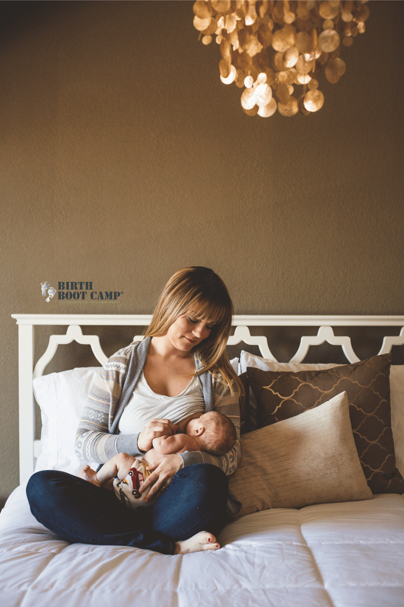 A woman is sitting on a bed with neutral-colored pillows, breastfeeding a baby. The room has a beige wall, a chandelier hanging from the ceiling, and a headboard with a white lattice design. There is a logo in the bottom left corner that reads 'Birth Boot Camp'.