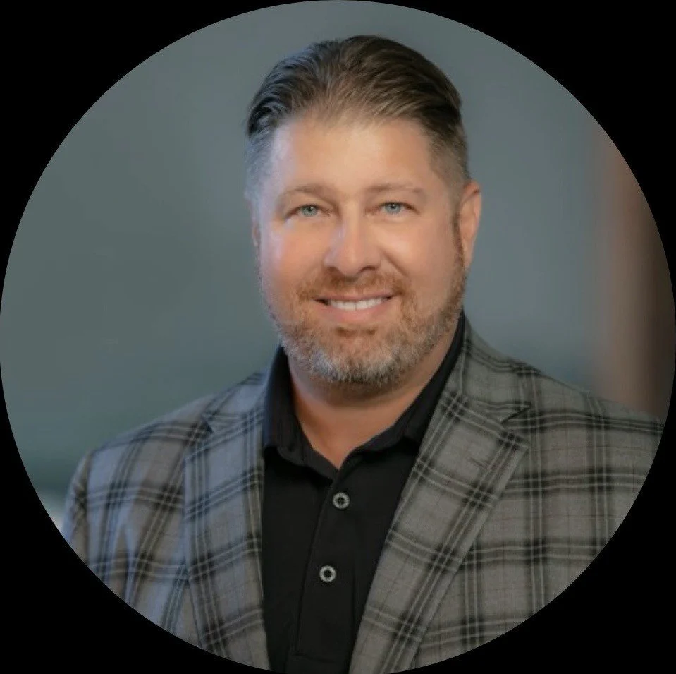 A professional headshot of a middle-aged man with short, light brown hair and a beard, wearing a gray plaid blazer over a black shirt, smiling at the camera against a blurred background.
