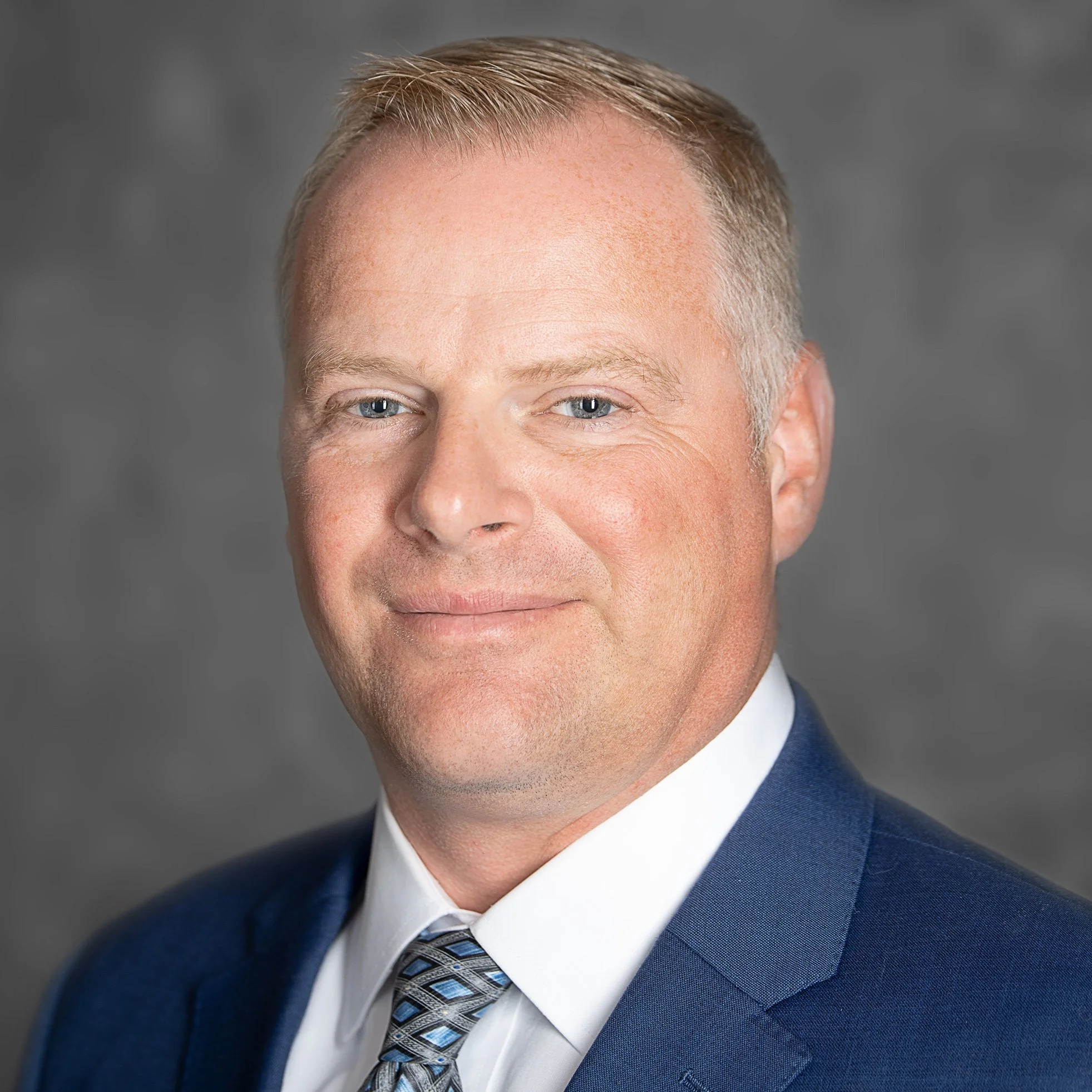 Close-up of a middle-aged man with blue eyes and short, light brown hair, wearing a blue suit, white shirt, and a patterned tie, smiling slightly against a gray background.
