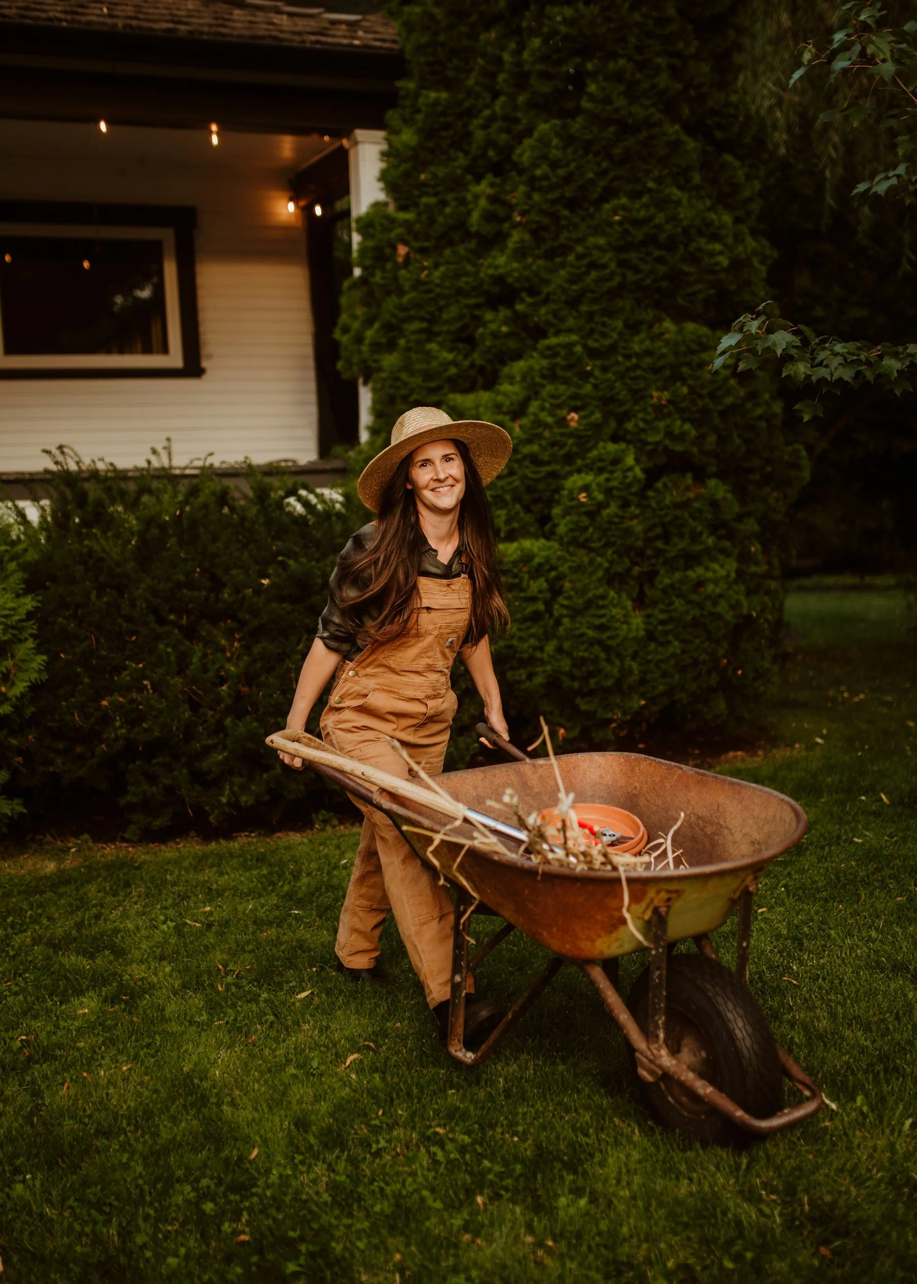 Woman smiling in garden pushing a rusty wheelbarrow filled with gardening debris, wearing a wide-brimmed hat and overalls.
