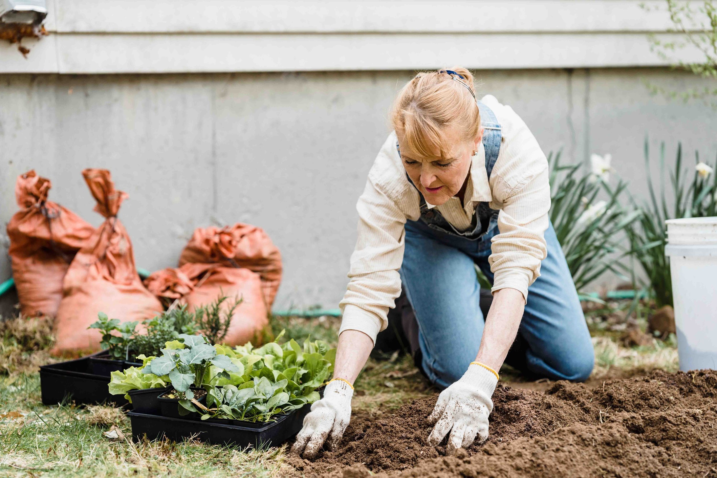 Woman planting vegetables in a garden, surrounded by gardening supplies and bags of soil.