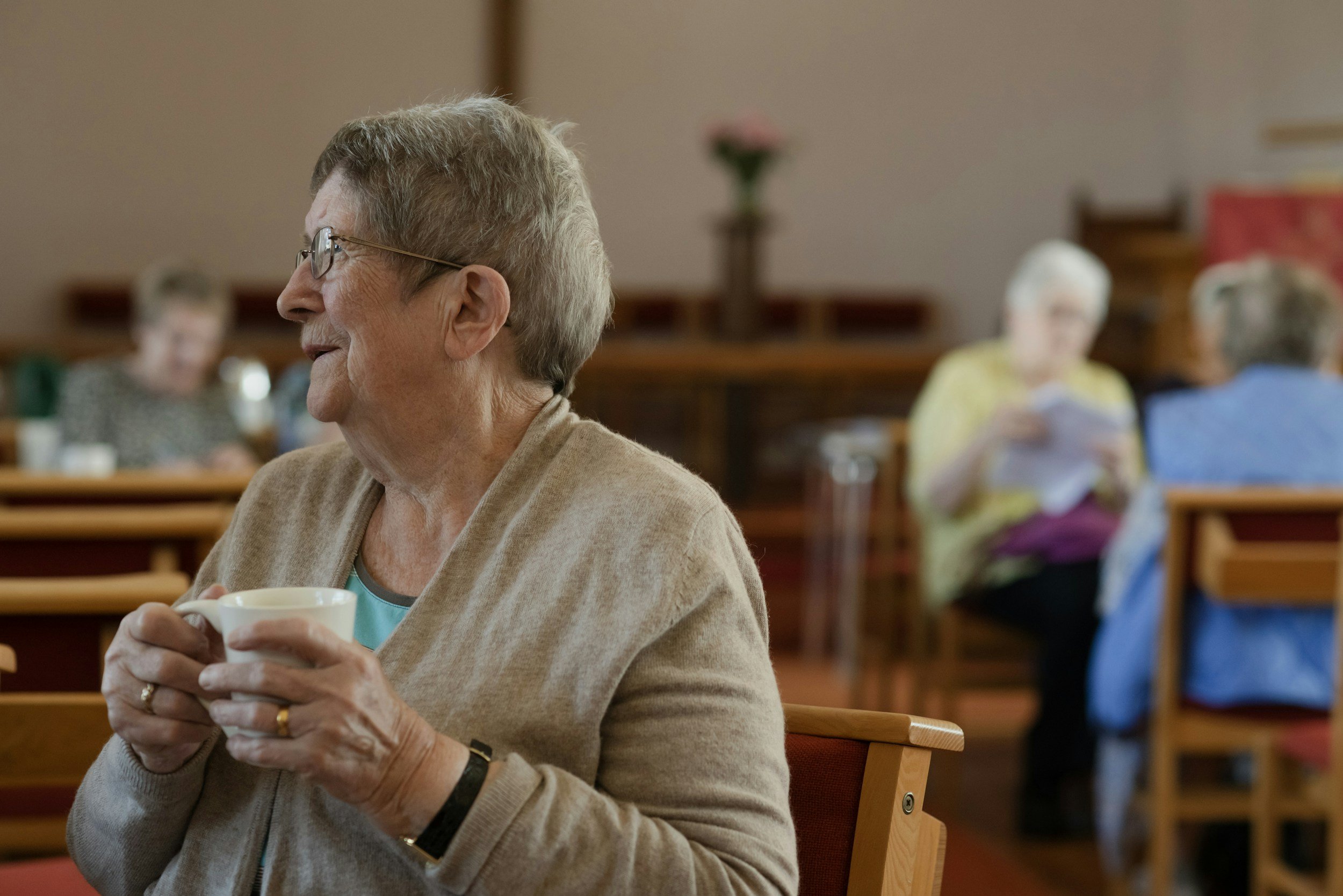 Elderly woman with glasses holding a cup, sitting at a table in a communal setting with other seniors in the background.