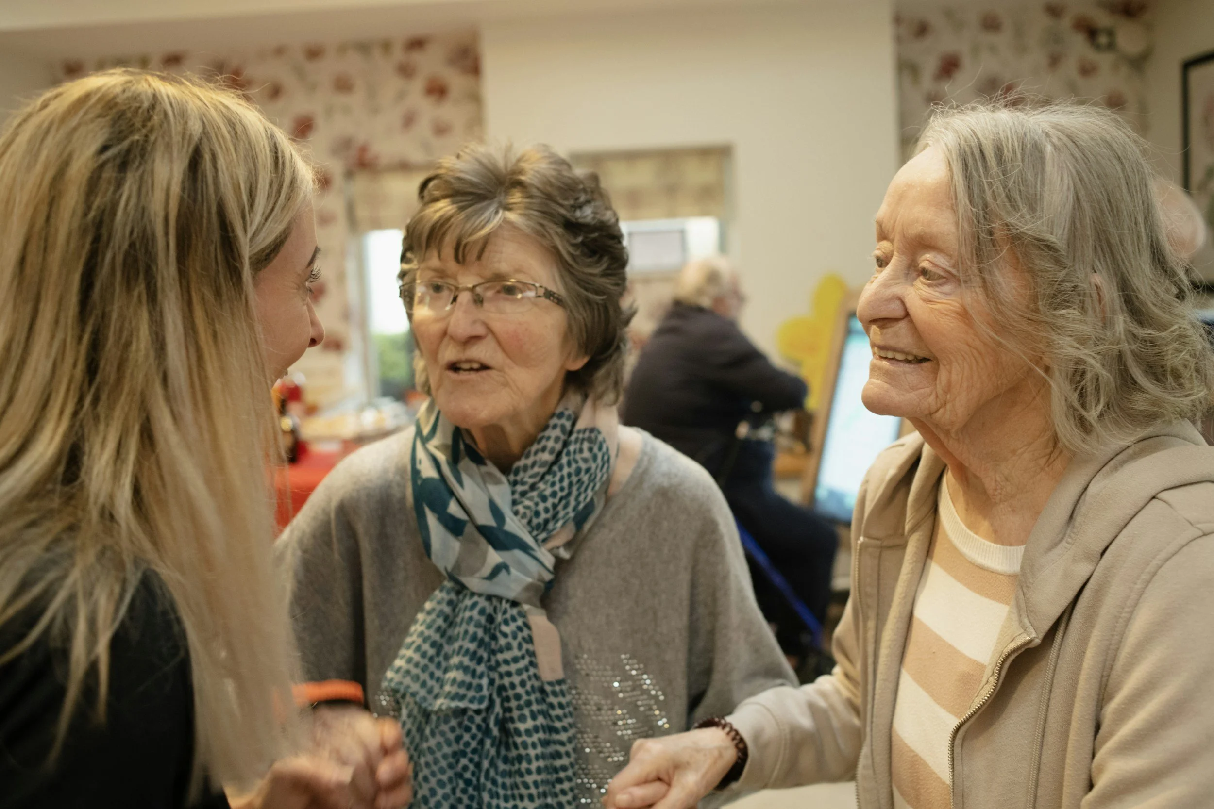 Three women engaged in conversation at an indoor gathering, with other people in the background.