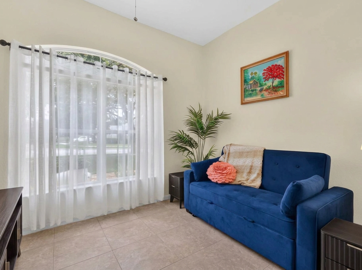 Living room with large window, white curtains, blue sofa with pink pillow, blanket, and bolster, potted plant, painting of a house and trees on wall.